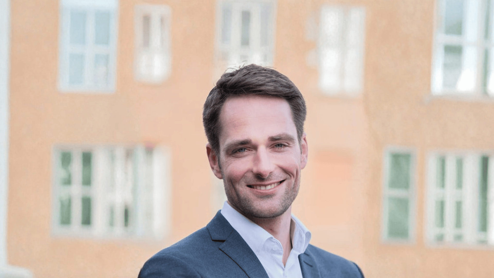 Ein Mann im Anzug steht im Freien und lächelt, während im unscharfen Hintergrund ein Gebäude mit großen Fenstern zu sehen ist.