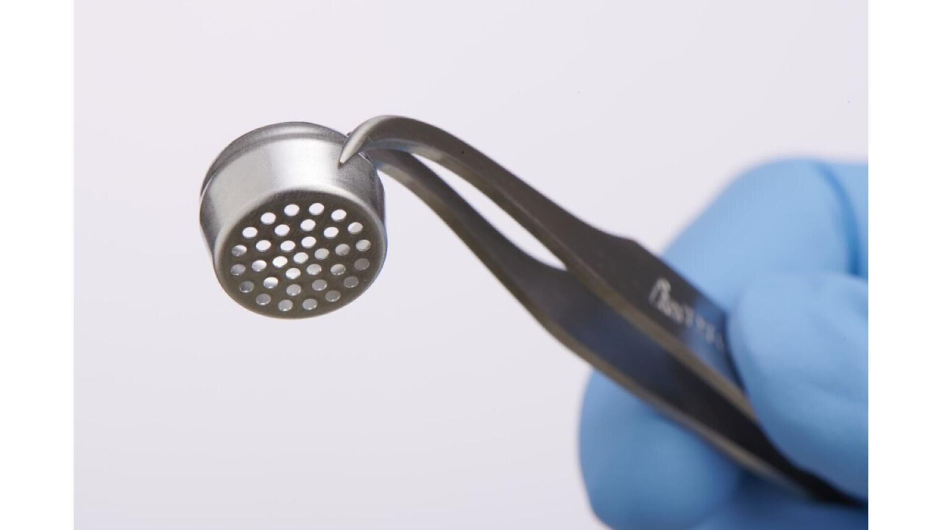 A gloved hand holds tweezers gripping a round metal object with multiple small holes on its flat surface, against a plain light background.