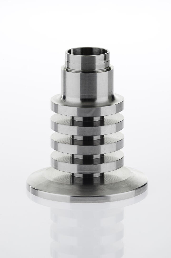 A stainless steel industrial metal fitting with a flanged base and a series of horizontal grooves around the cylindrical body, placed on a reflective white surface.