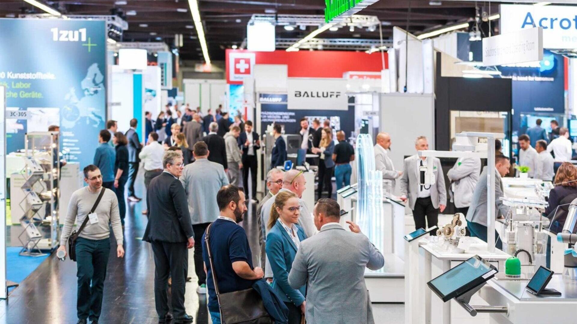 A busy trade show floor with many people walking, talking, and visiting various technology and industrial booths, surrounded by large banners, displays, and digital screens.