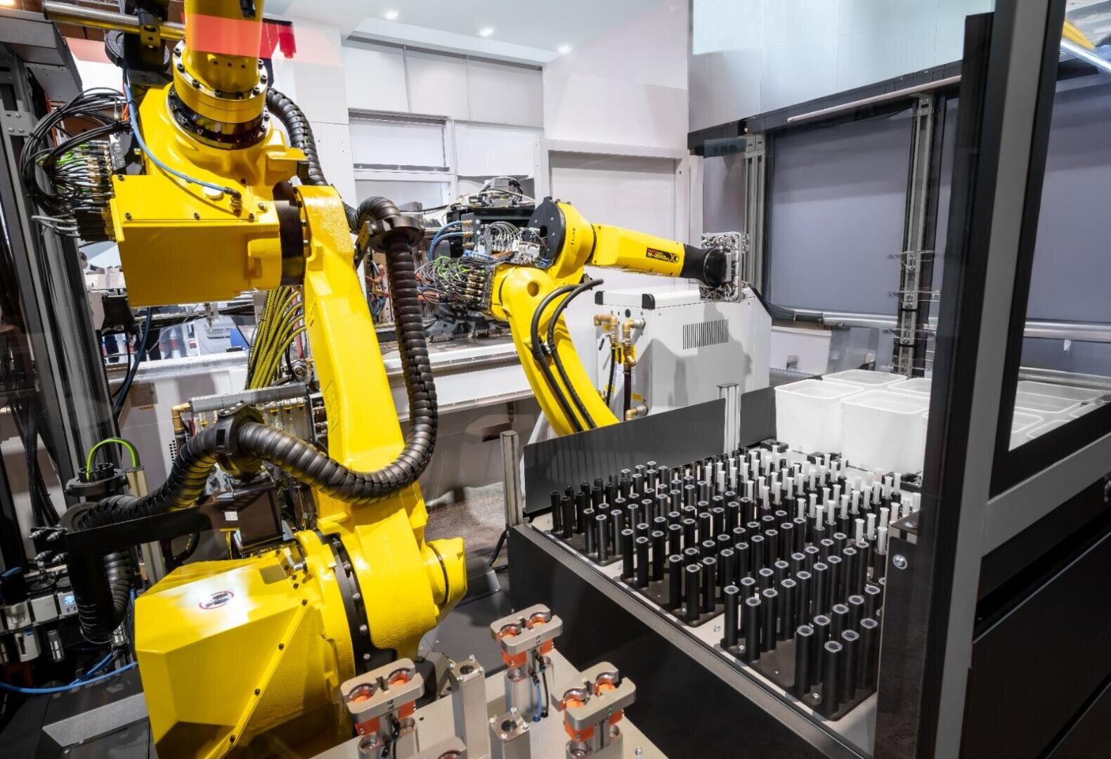 Zwei gelbe Industrieroboterarme arbeiten in einer automatisierten Fertigungsanlage und handhaben zylindrische Metallteile auf einer Fertigungsstraße, die von Maschinen und Lagerbehältern umgeben ist.