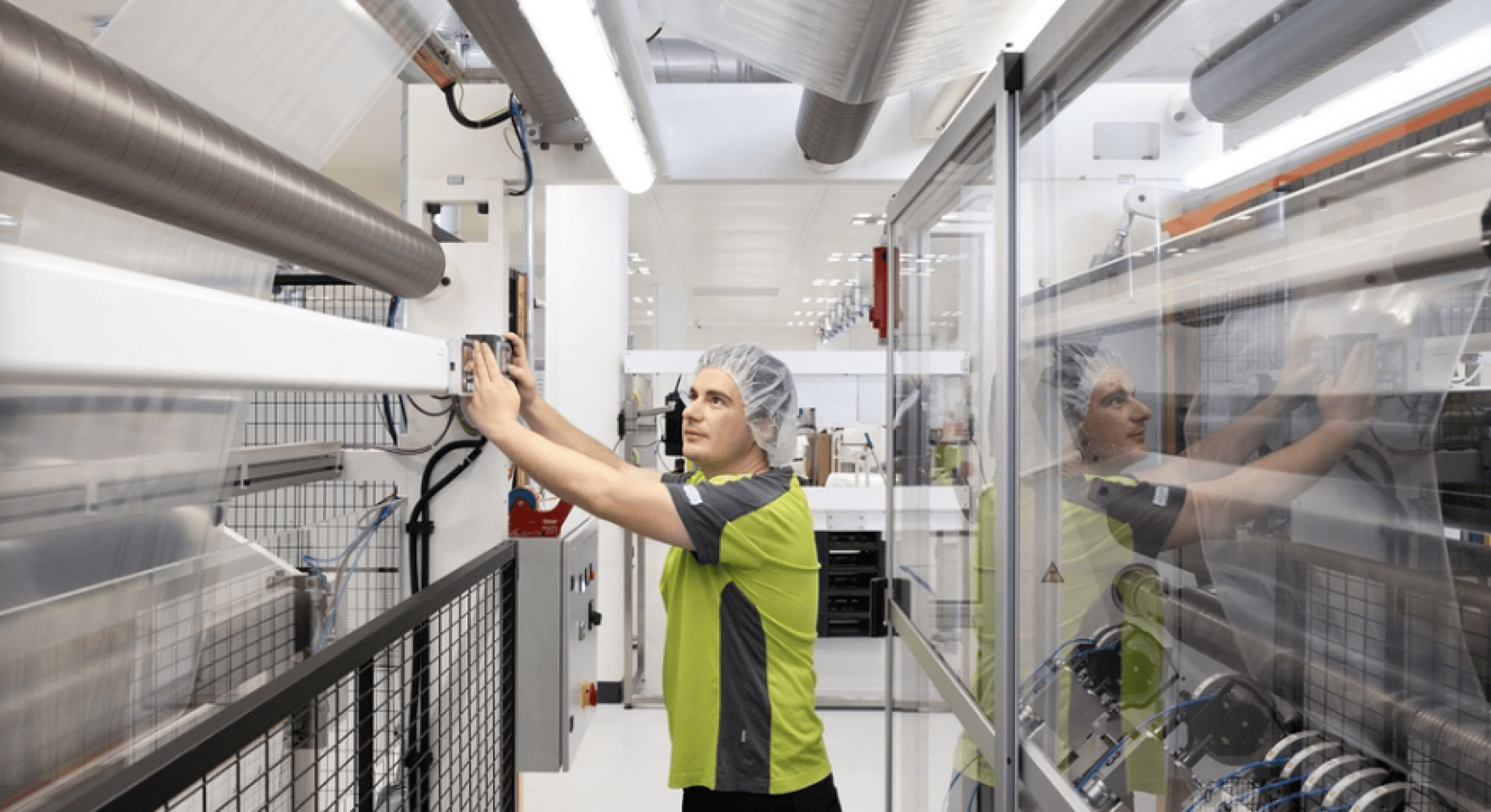 A person wearing a hair net and a green-and-gray uniform operates machinery in a clean, modern industrial facility, surrounded by metal pipes, equipment, and glass panels.