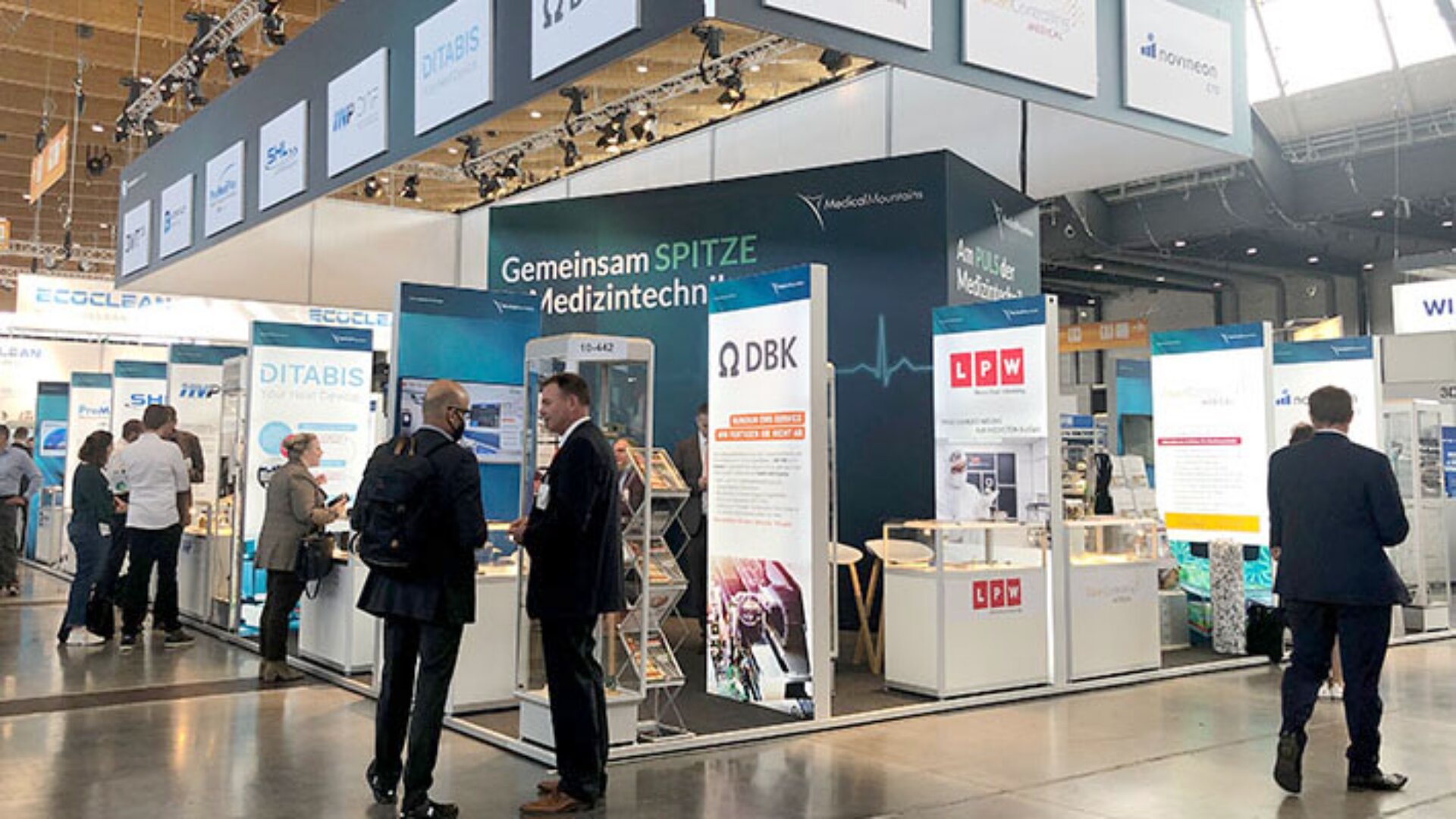 Eine geschäftige Ausstellungshalle mit Menschen, die sich an Ständen mit medizintechnischen Produkten und Bannern unter einem großen Schild mit der Aufschrift „Gemeinsam SPITZE in Medizintechnik“ unterhalten.