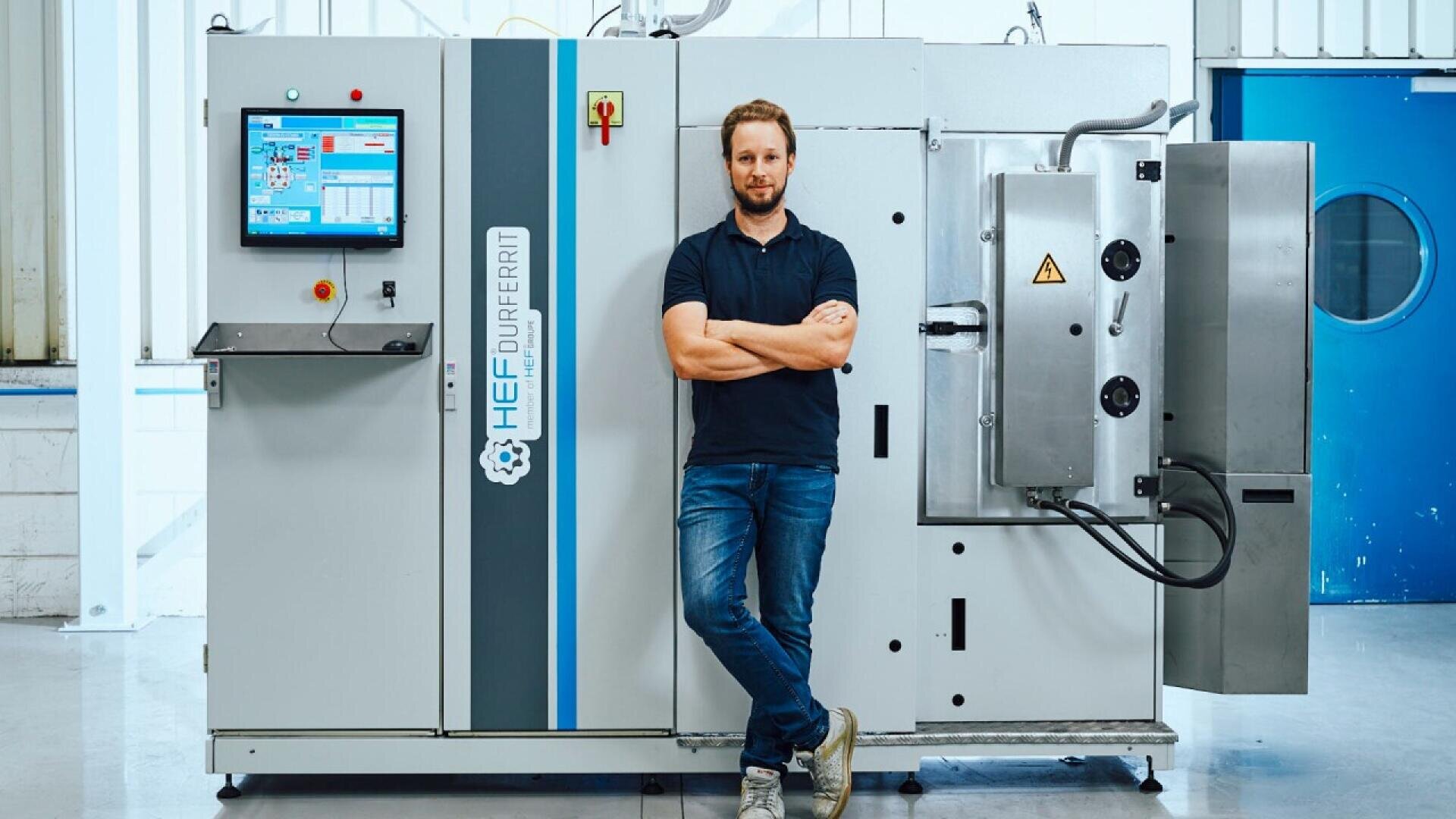 A man in a navy shirt and jeans stands with arms crossed in front of a large industrial machine with a touchscreen control panel in a clean, modern facility.