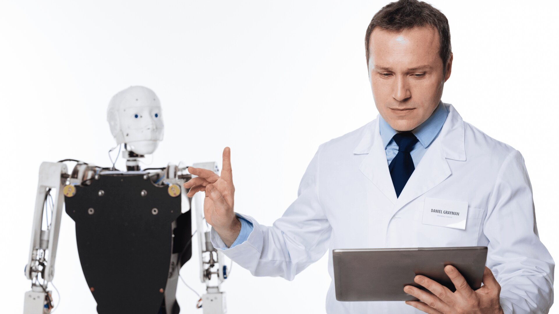 Ein Wissenschaftler im weißen Laborkittel hält ein Tablet und gestikuliert mit der Hand. Neben ihm steht ein humanoider Roboter mit weißem Gesicht und mechanischem Körper. Der Hintergrund ist schlicht weiß.