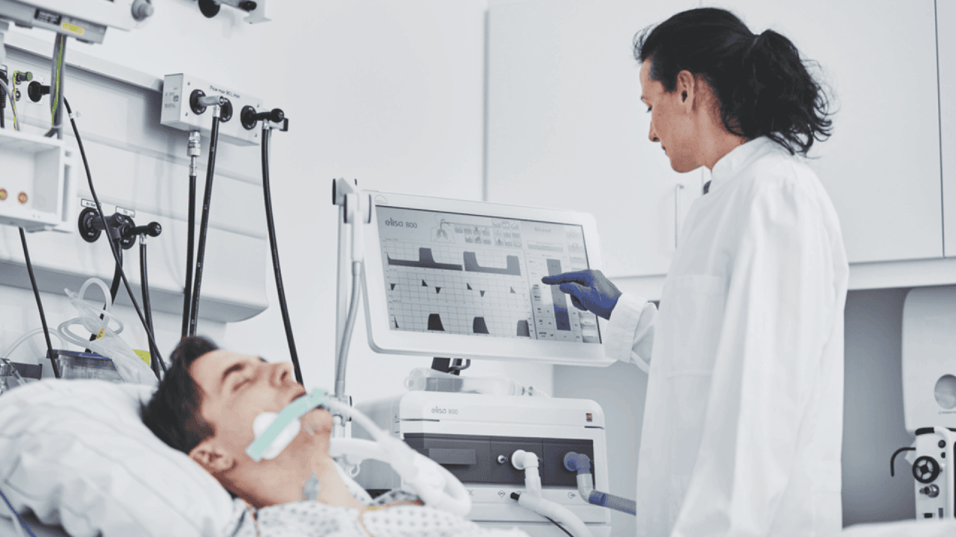 A healthcare worker in a lab coat and gloves monitors a patient on a ventilator, interacting with a touchscreen medical monitor in a hospital room. The patient lies in bed with a breathing tube.