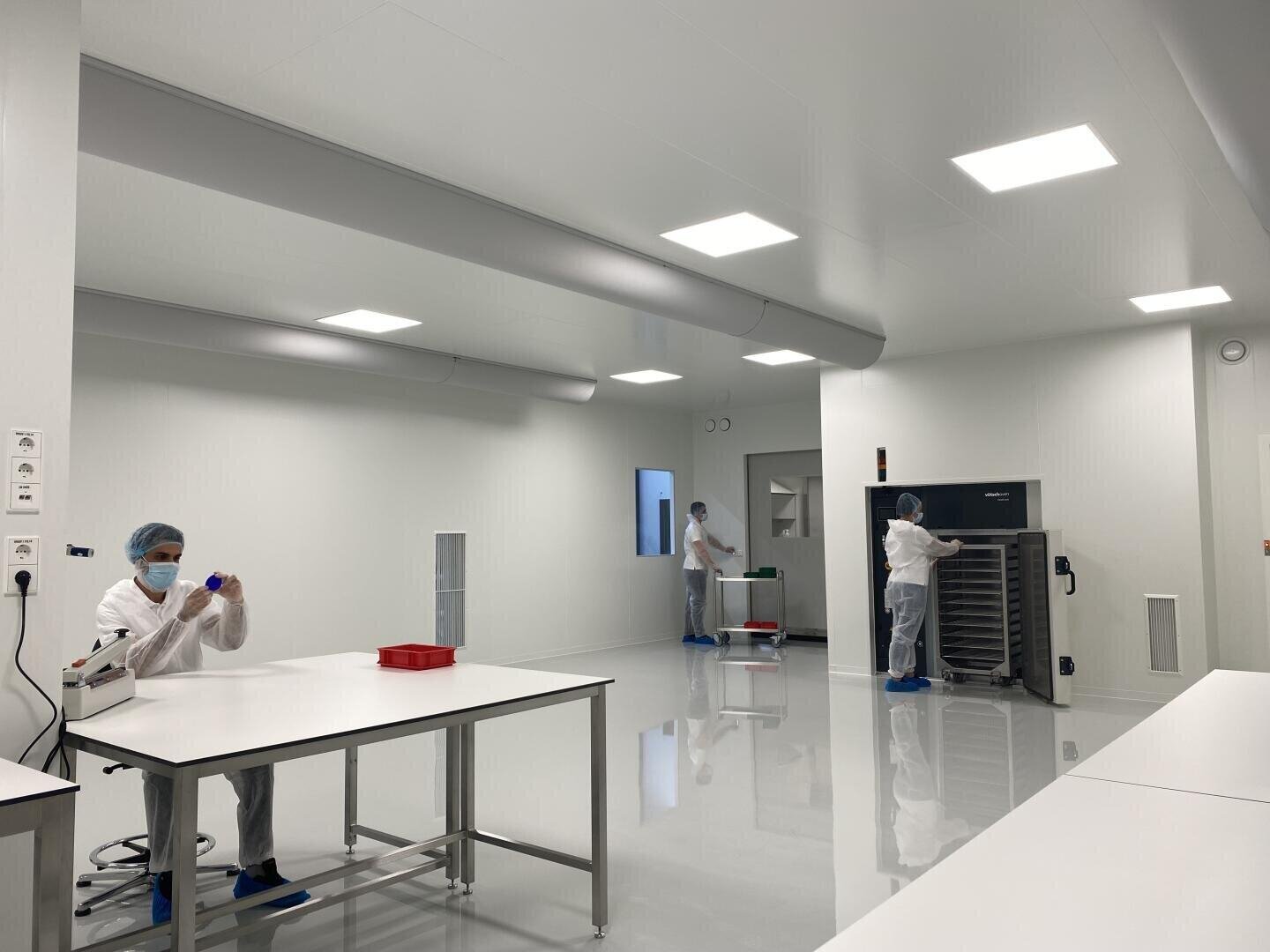 Three people in white lab coats, masks and hairnets work in a clean, bright laboratory with white walls, shiny floors and stainless steel equipment. One sits at a table while two stand next to a large machine.