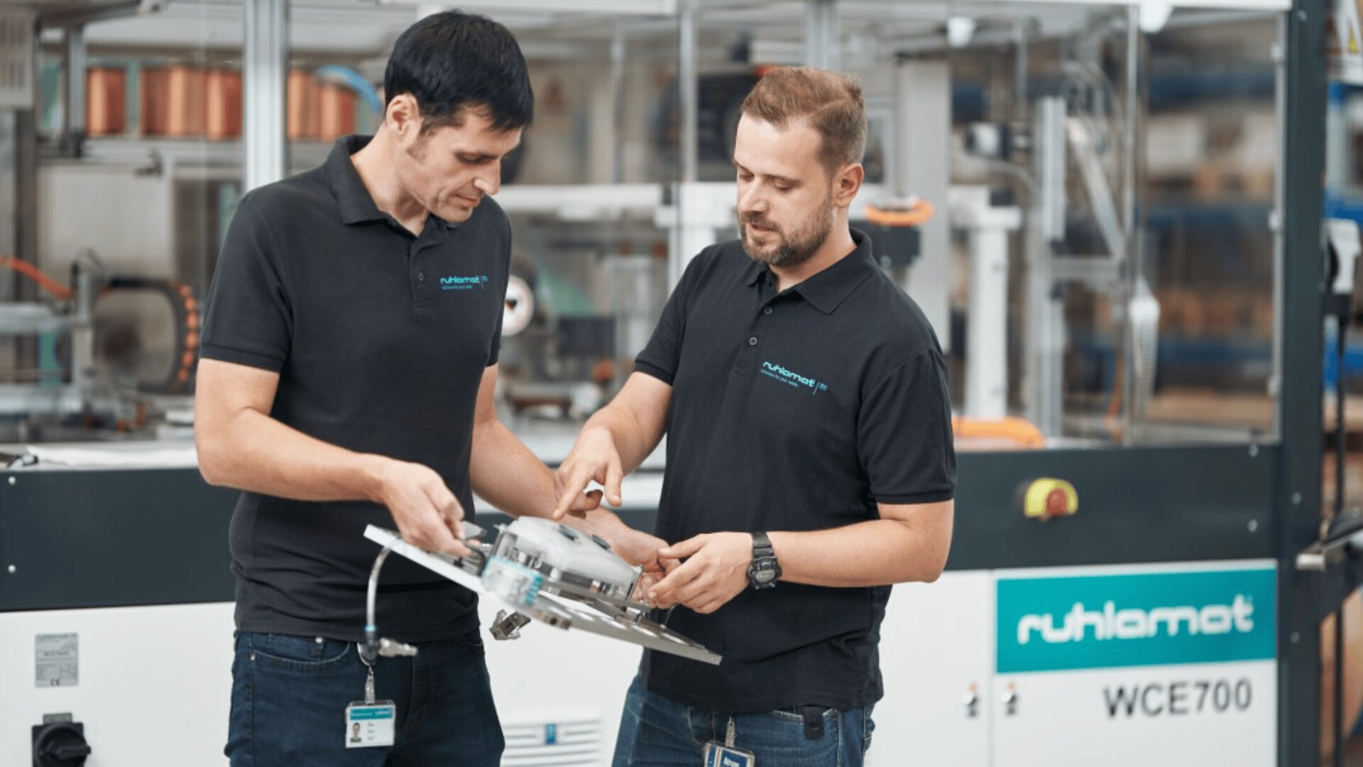 Two men wearing black polo shirts stand in a factory, examining and discussing a circular metal component. Machinery and equipment are visible in the background, including one labeled ruthlamat WCE700.