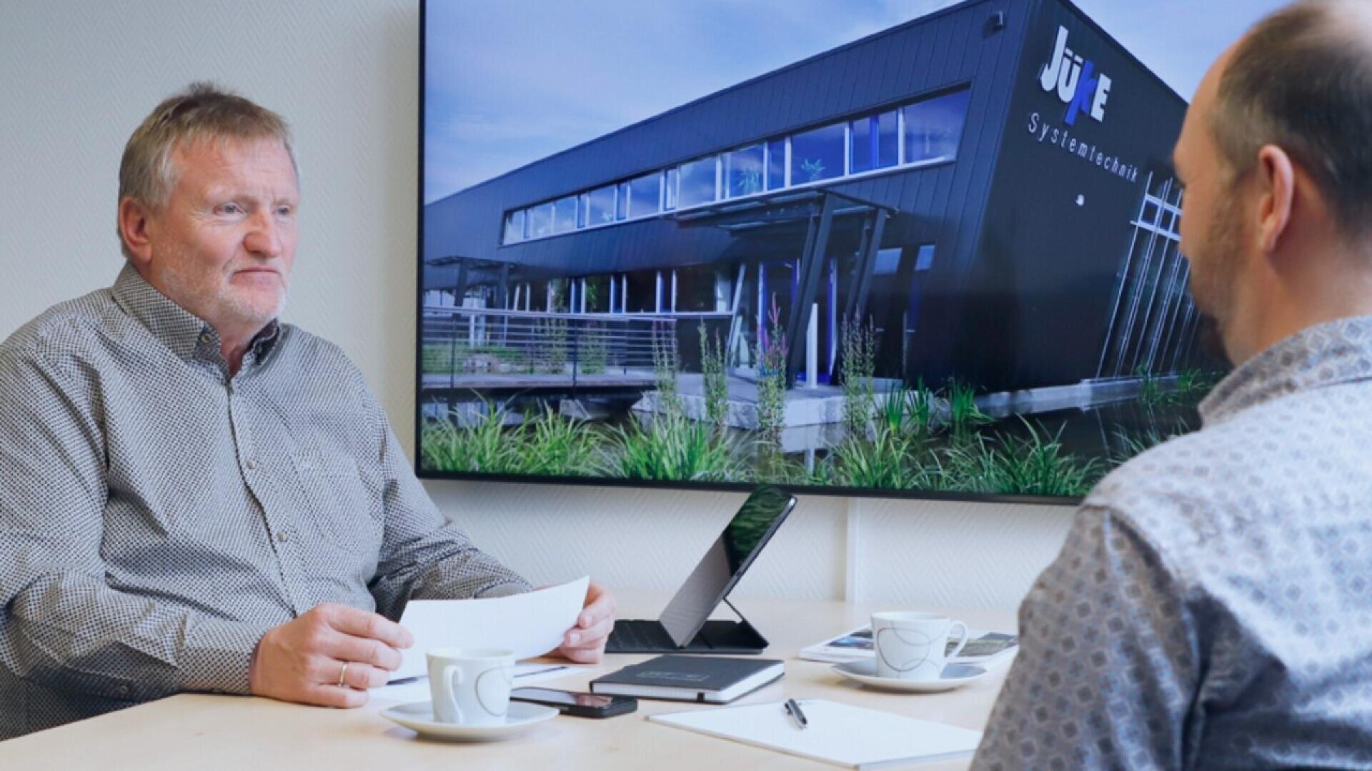 Two men sit at a table with papers, coffee cups, and a laptop. A large screen behind them displays the exterior of a modern building with the sign “JUIE Systemtechnik.”.