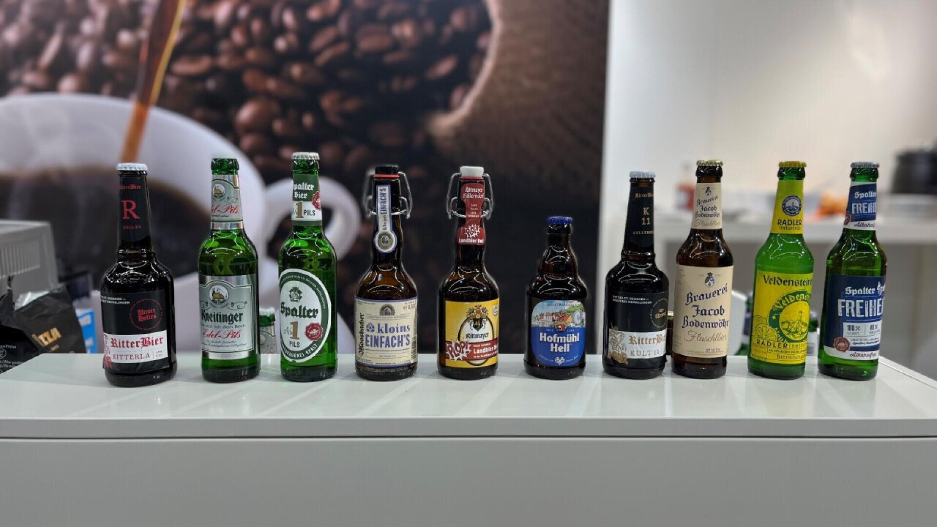 Ten assorted German beer bottles with different labels and colors are lined up on a white surface, with a blurred background featuring a coffee beans image and white walls.