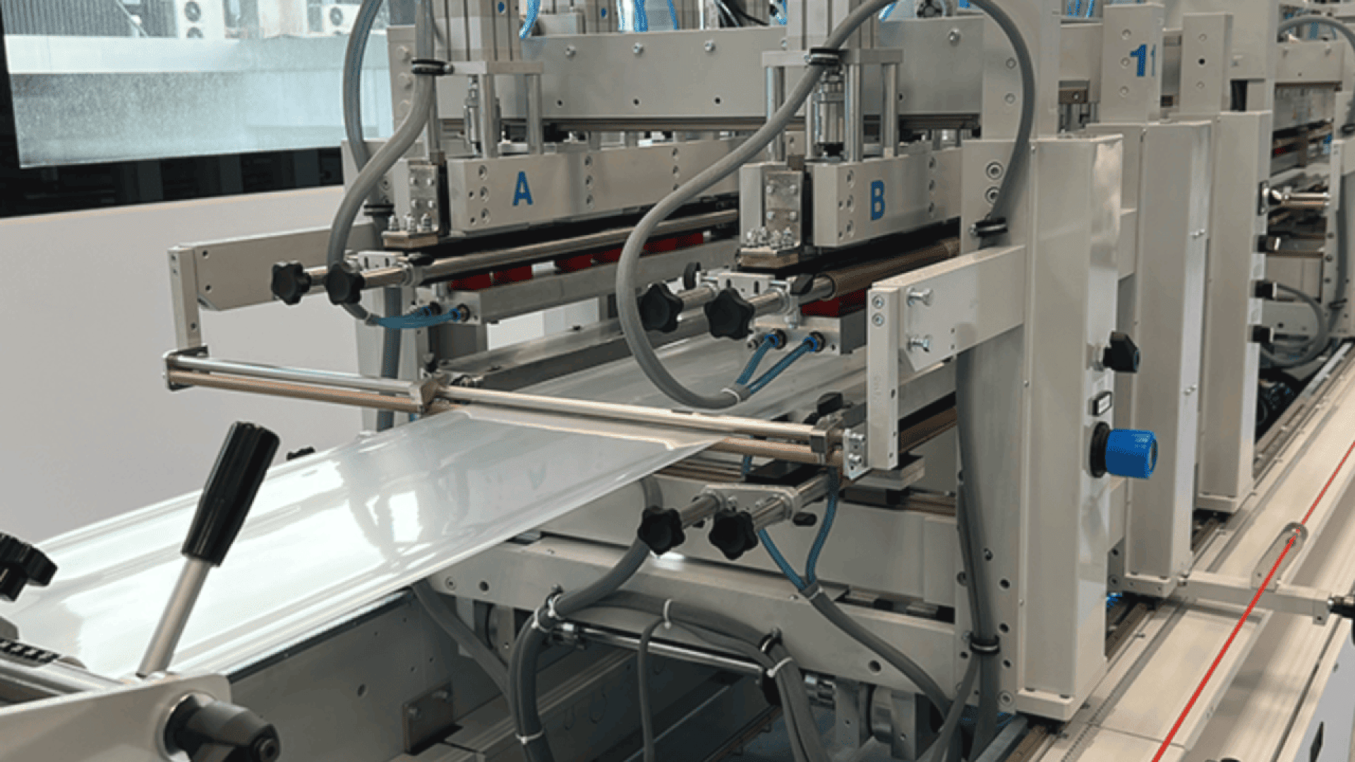 Close-up of an automated industrial packaging machine with robotic arms and rollers handling a sheet of plastic film in a factory setting.