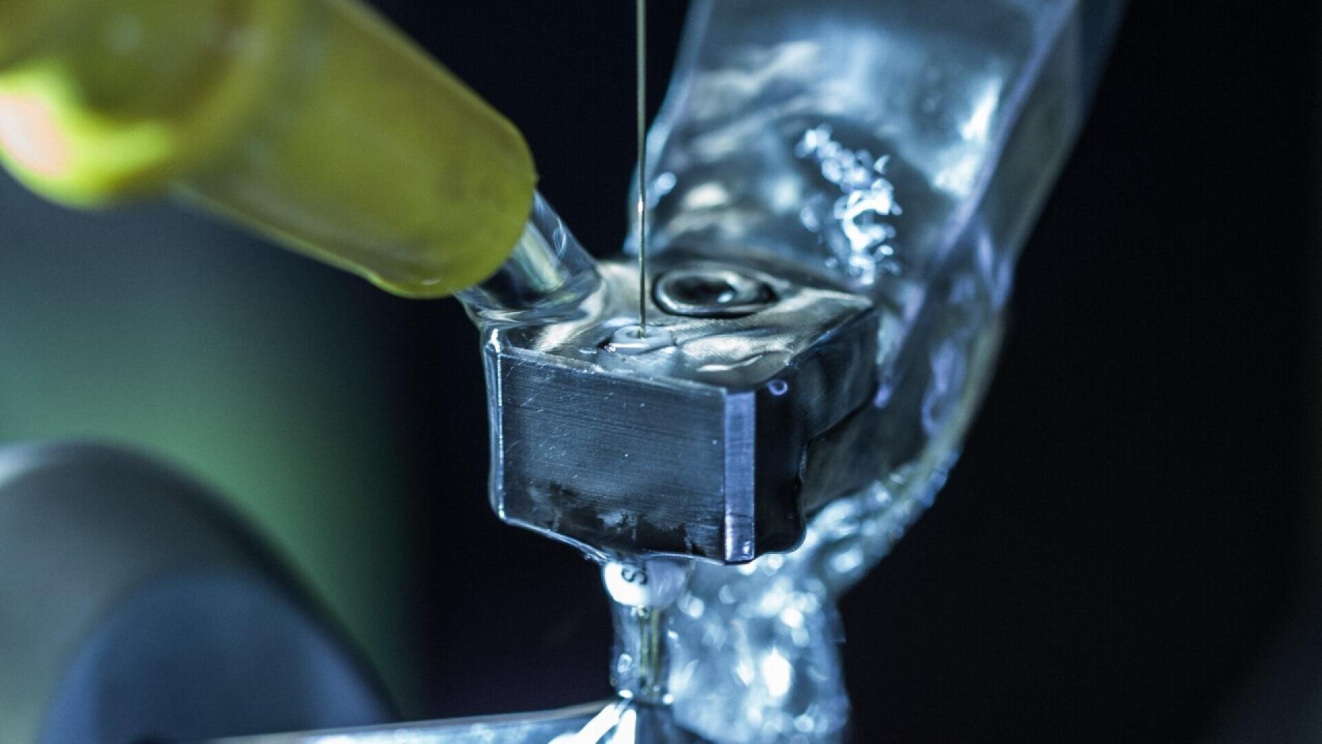 Close-up of a metalworking process with a needle or thin nozzle injecting liquid onto a shiny metal component, likely for cooling or lubrication, with a blurred background.
