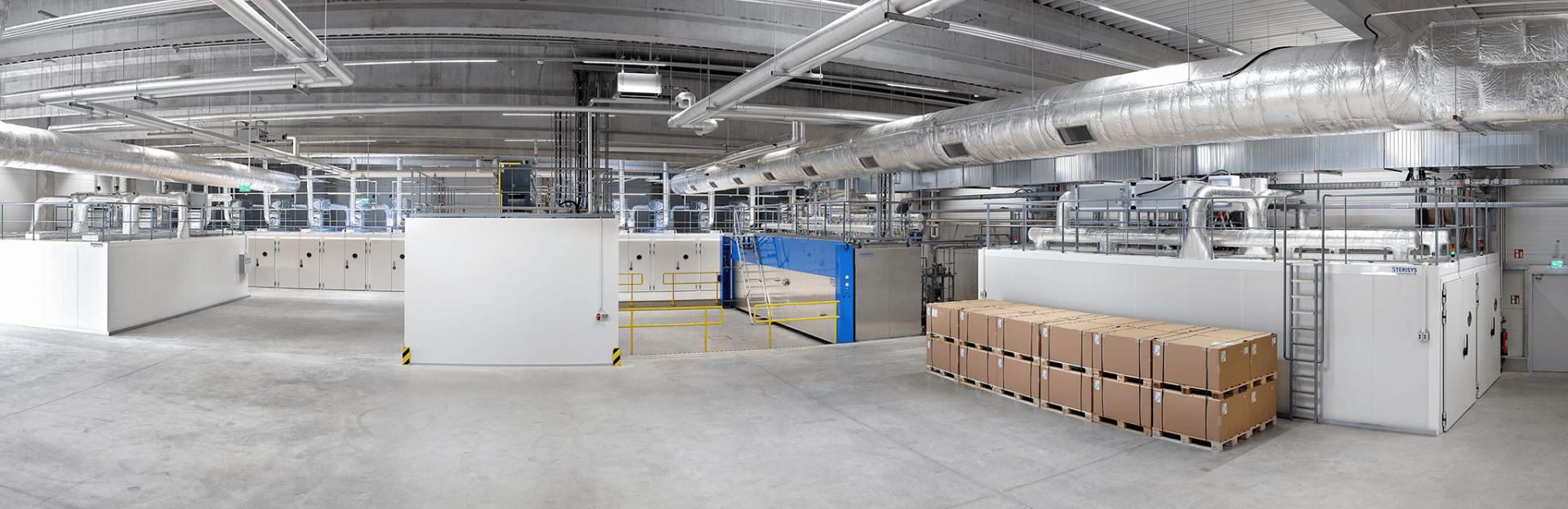 Eine geräumige Industrieanlage mit weißen Wänden, großen Lüftungsschächten und gestapelten Kartons auf Paletten; der Raum ist sauber und gut beleuchtet und hat einen Betonboden.