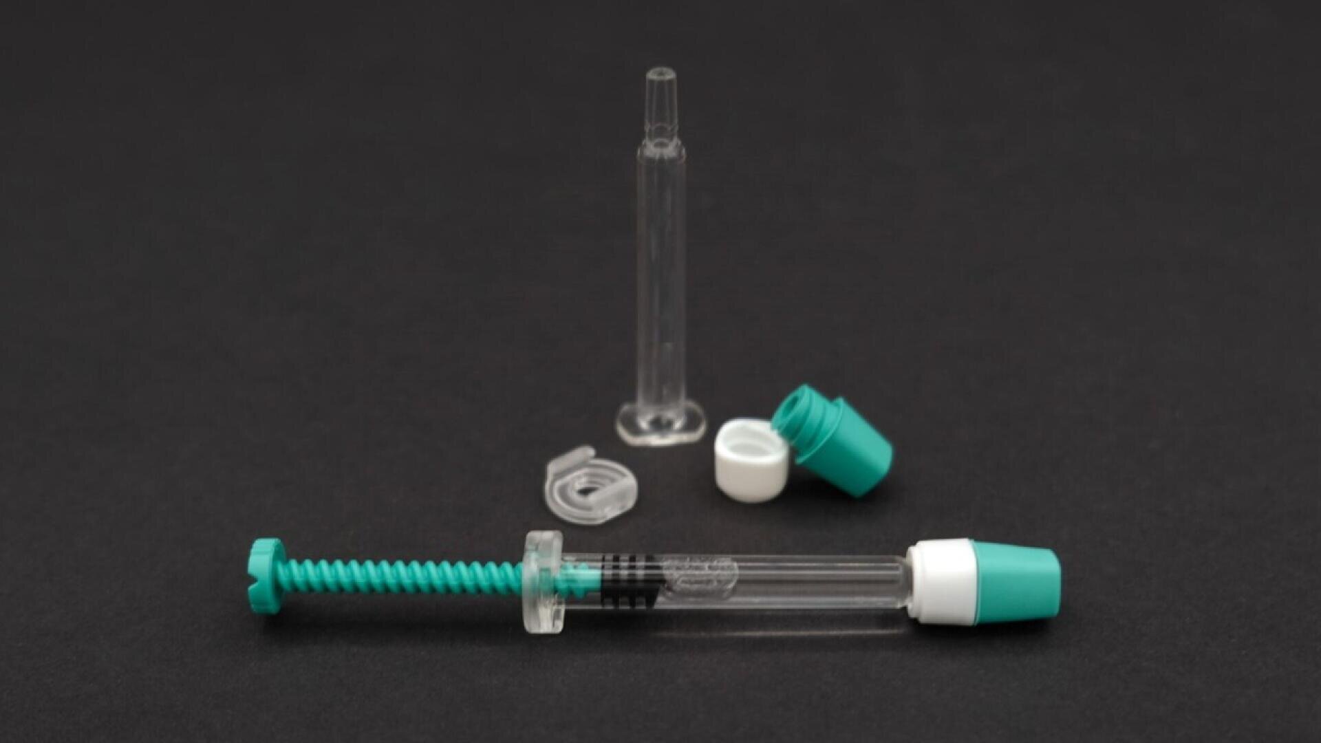 A clear plastic dental syringe with a green plunger and cap, shown disassembled with additional clear and green components, displayed on a plain black background.