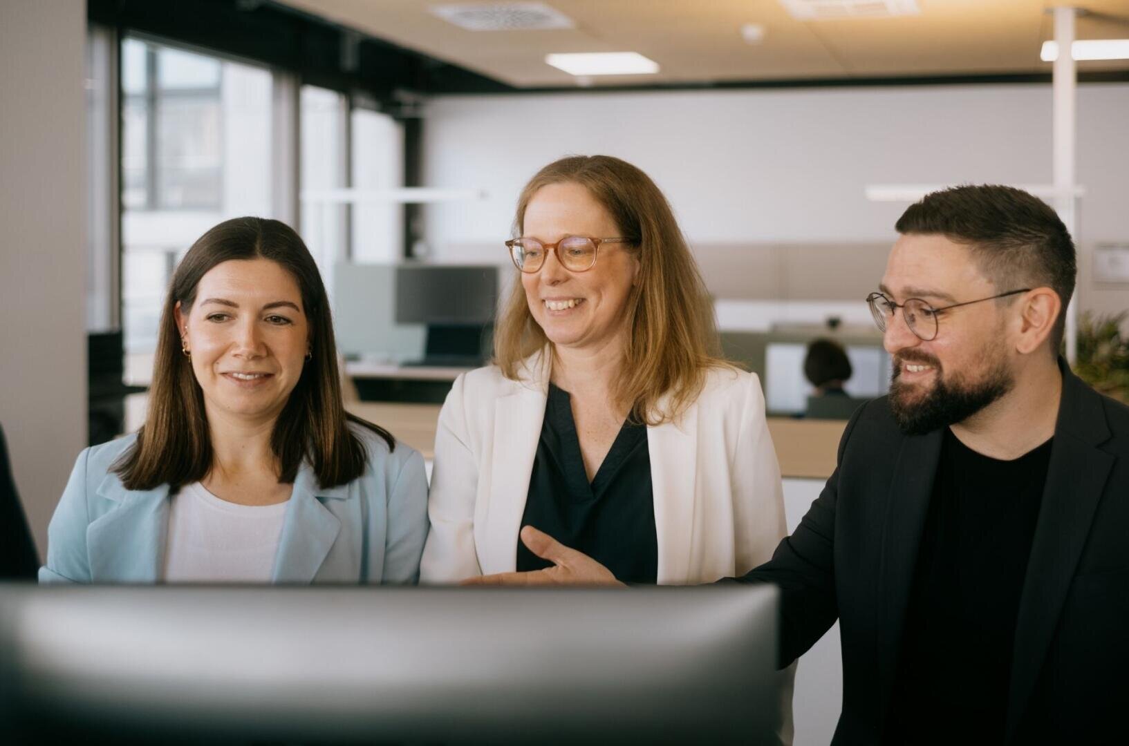 Three professionally dressed people, two women and a man, are smiling and looking at a computer screen in a modern office environment. It looks as if they are working together or discussing something together.
