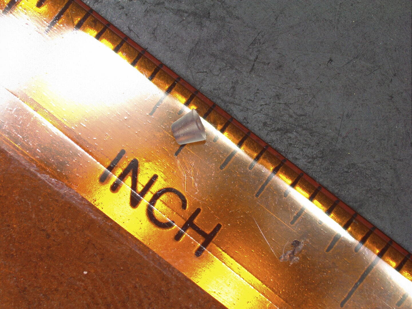 Ein winziges, metallisches, kegelförmiges Objekt wird auf einem Lineal neben der Zoll-Markierung platziert. Dies zeigt seine geringe Größe von weniger als einem halben Zoll Länge. Die Hintergrundoberfläche ist dunkel und strukturiert.