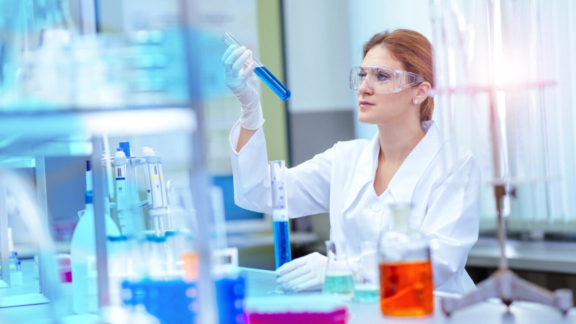 A scientist in a white lab coat and safety goggles examines a blue liquid in a test tube in a laboratory filled with various glassware containing colorful liquids.