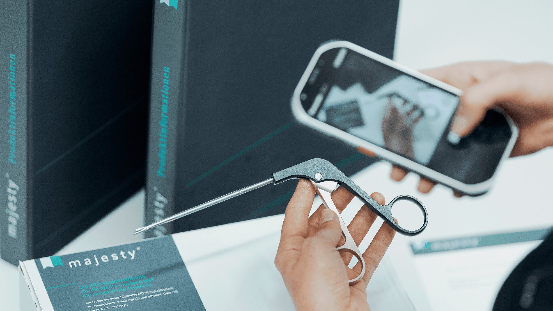 Eine Person hält ein medizinisches Instrument in einer Hand und fotografiert mit der anderen Hand ein Smartphone des Instruments. Im Hintergrund sind Markenboxen mit der Aufschrift „Majesty“ zu sehen.