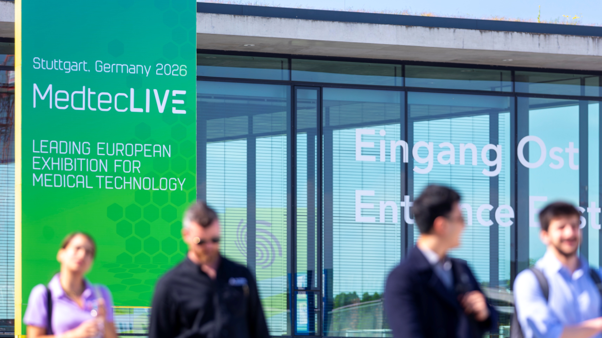 Large green sign for MedtecLIVE, a leading European medical technology exhibition in Stuttgart, Germany 2026, is displayed outside a glass building with blurred people walking in the foreground.