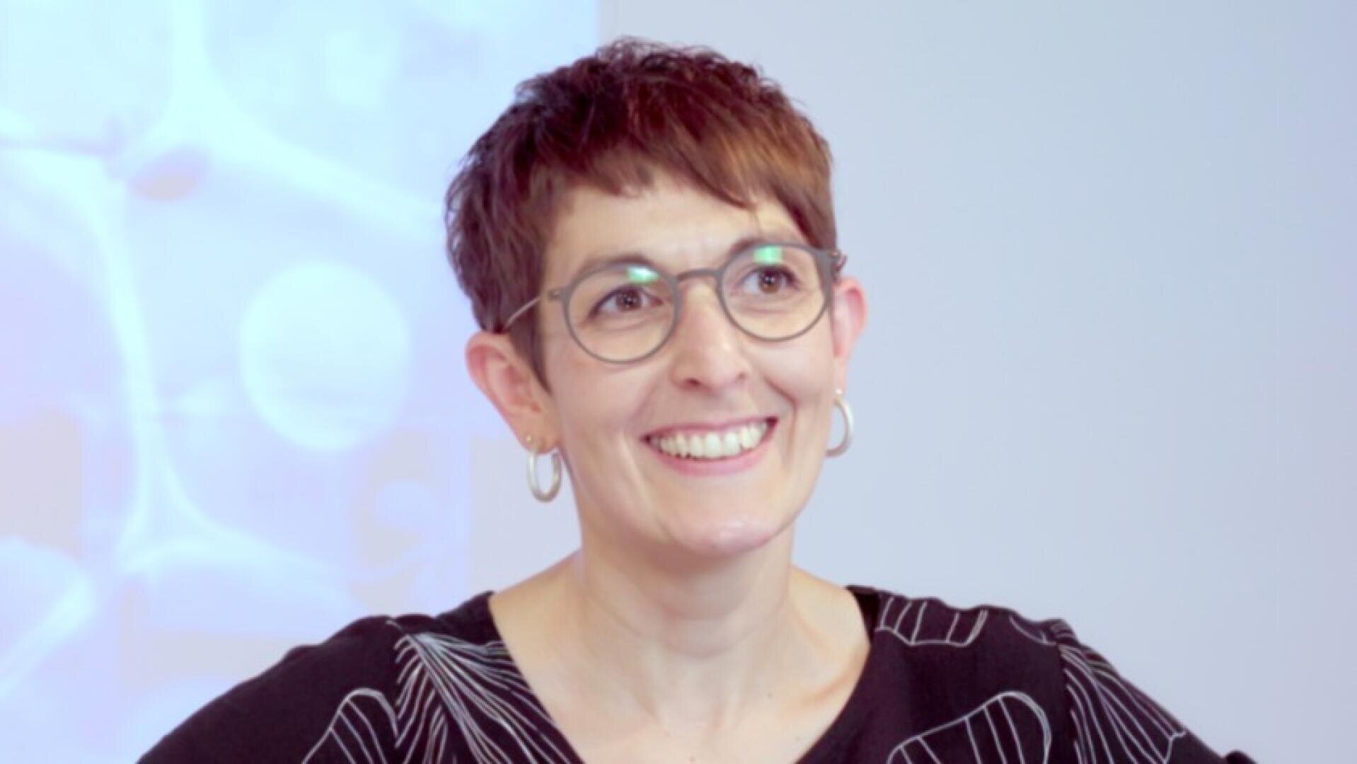 Eine Frau mit kurzen braunen Haaren, Brille und Ohrringen lächelt, während sie leicht aus der Kamera schaut. Sie trägt ein schwarzes Oberteil mit weißem Muster, und der Hintergrund ist sanft verschwommen.