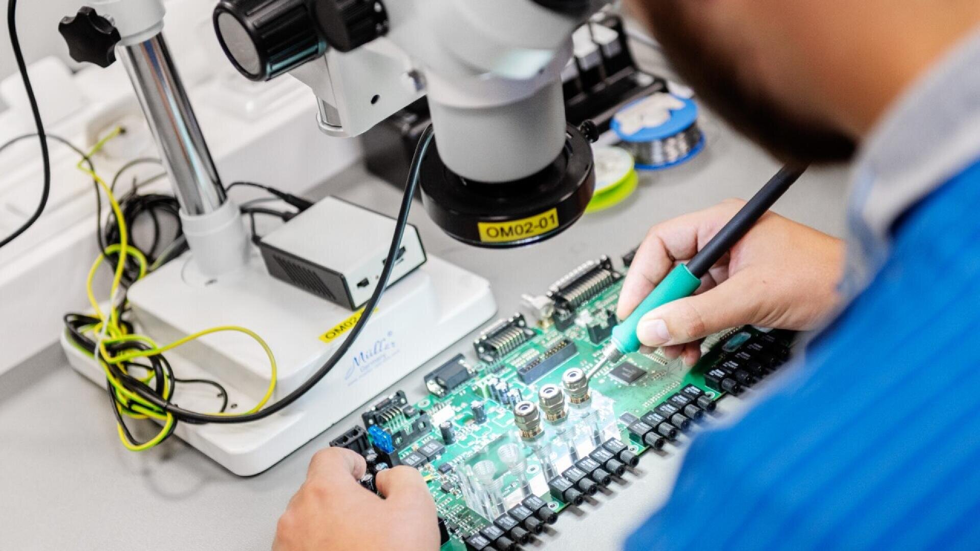A person uses a soldering tool to work on a green circuit board under a microscope in a laboratory setting, focusing on intricate electronic components.