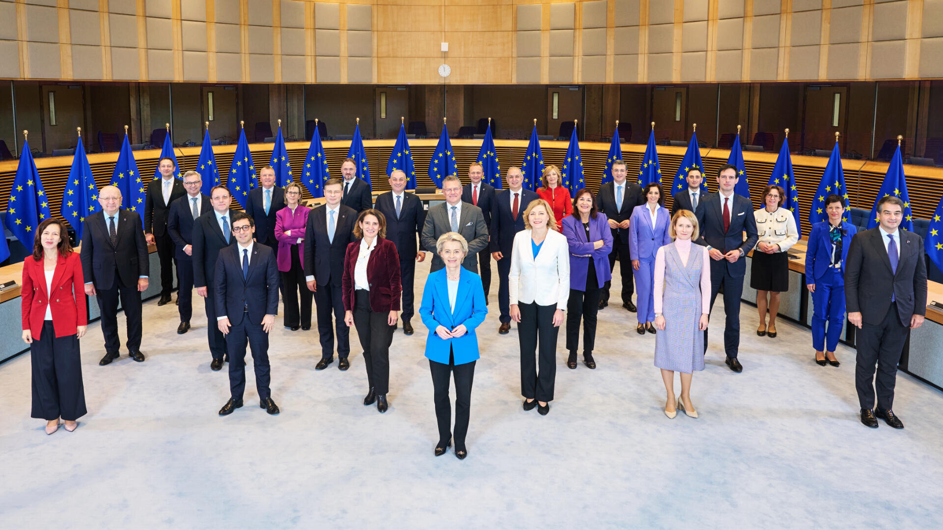 Eine Gruppe formell gekleideter Beamter, darunter Männer und Frauen, steht mit Abstand in einem großen Raum, hinter ihnen mehrere EU-Flaggen, und posiert für ein Gruppenfoto.