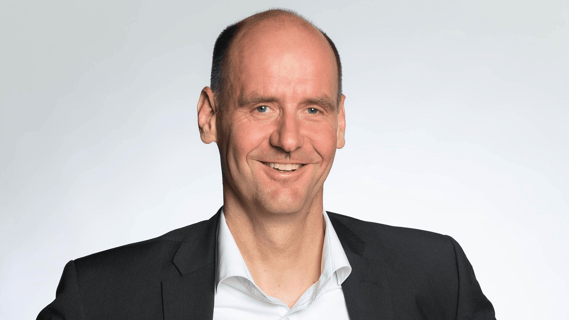 Ein Mann mittleren Alters mit zurückweichendem Haaransatz, der ein dunkles Jackett und ein weißes Hemd trägt, lächelt vor einem schlichten hellgrauen Hintergrund in die Kamera.