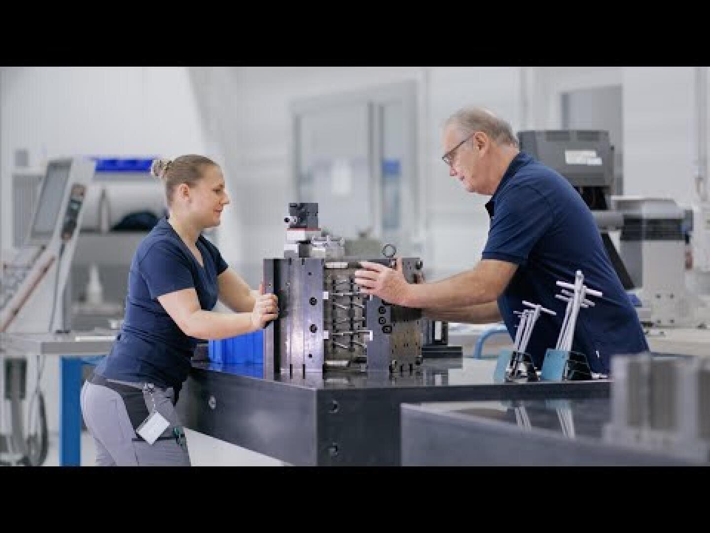 Zwei Personen in einer Werkstatt, beide in marineblauen Hemden, arbeiten gemeinsam an einem großen Maschinenteil aus Metall, das auf einer Werkbank liegt. Die Umgebung erscheint sauber und organisiert, mit verschiedenen Werkzeugen und Geräten im Hintergrund.