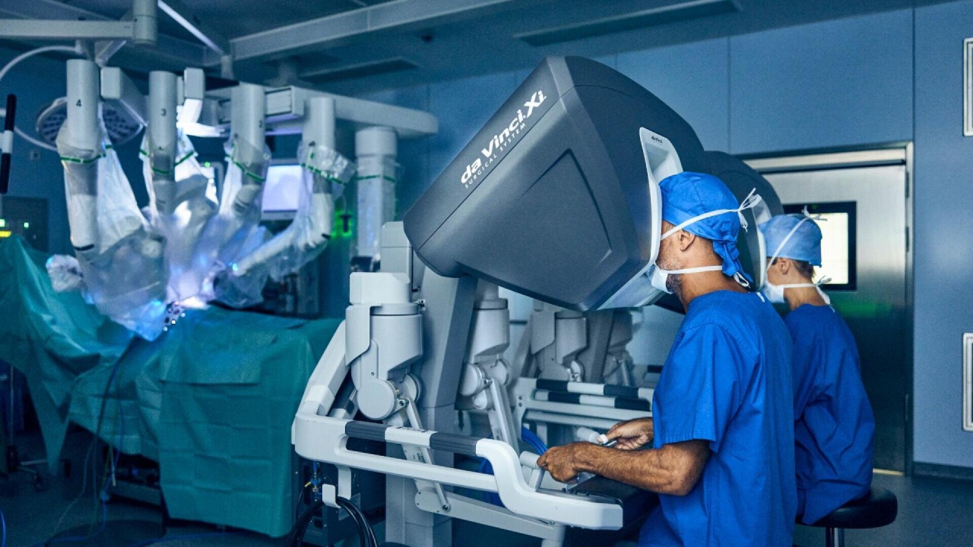 Two surgeons in blue scrubs operate a robotic surgical system in a modern operating room, looking into control consoles while robotic arms perform surgery on a patient in the background.
