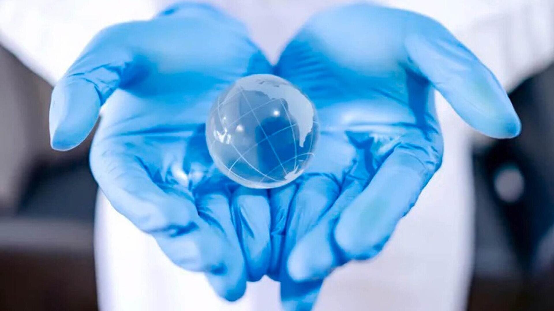 A person wearing blue gloves gently holds a small, transparent glass globe with map lines, symbolizing global care or protection, in their cupped hands.