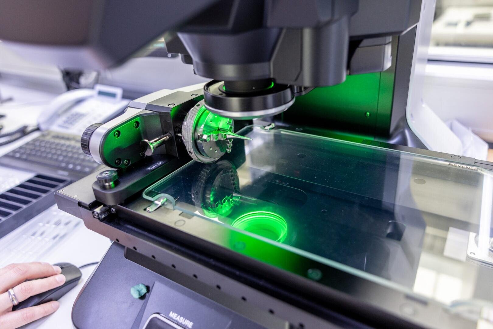 Nahaufnahme eines Hightech-Mikroskops mit einem Objektträger, der unter grünem Licht in einer Laborumgebung untersucht wird, wobei links die Hand einer Person teilweise sichtbar ist.