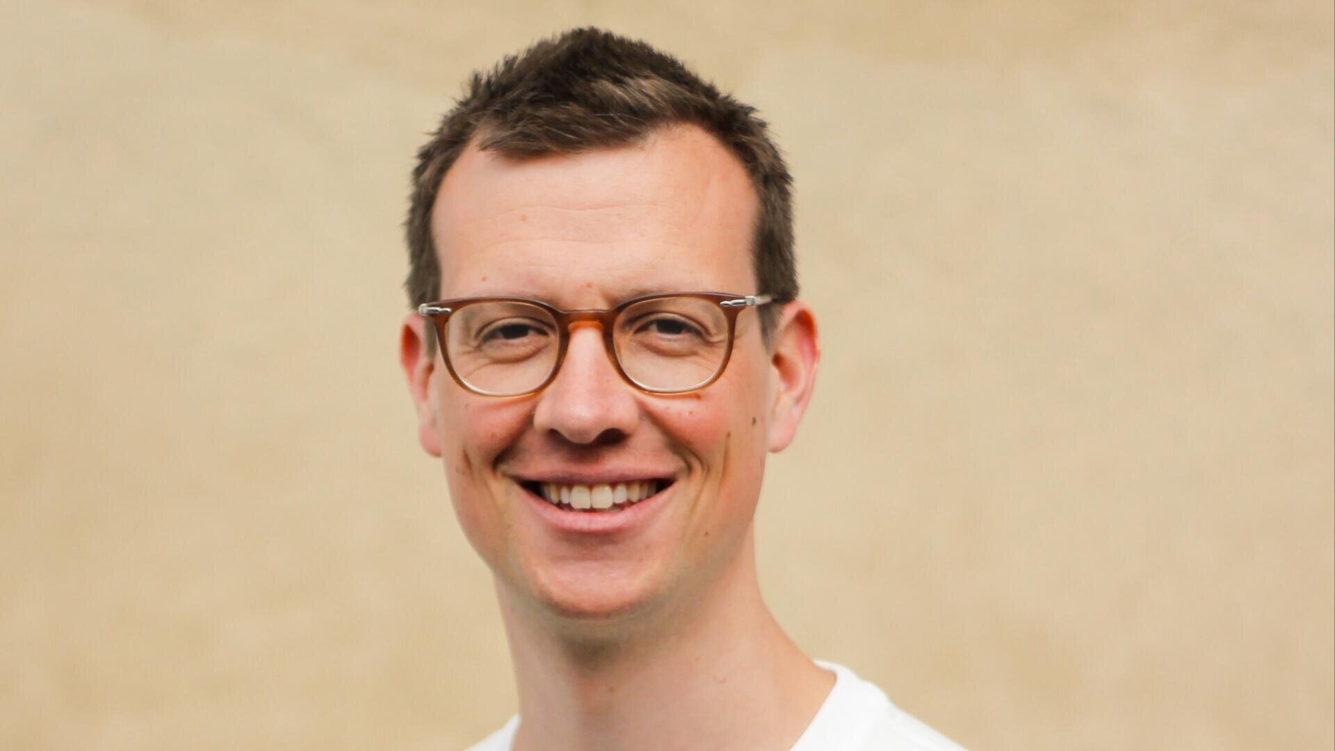 A man with short brown hair and glasses smiles at the camera. He is wearing a white shirt and standing in front of a plain, beige background.