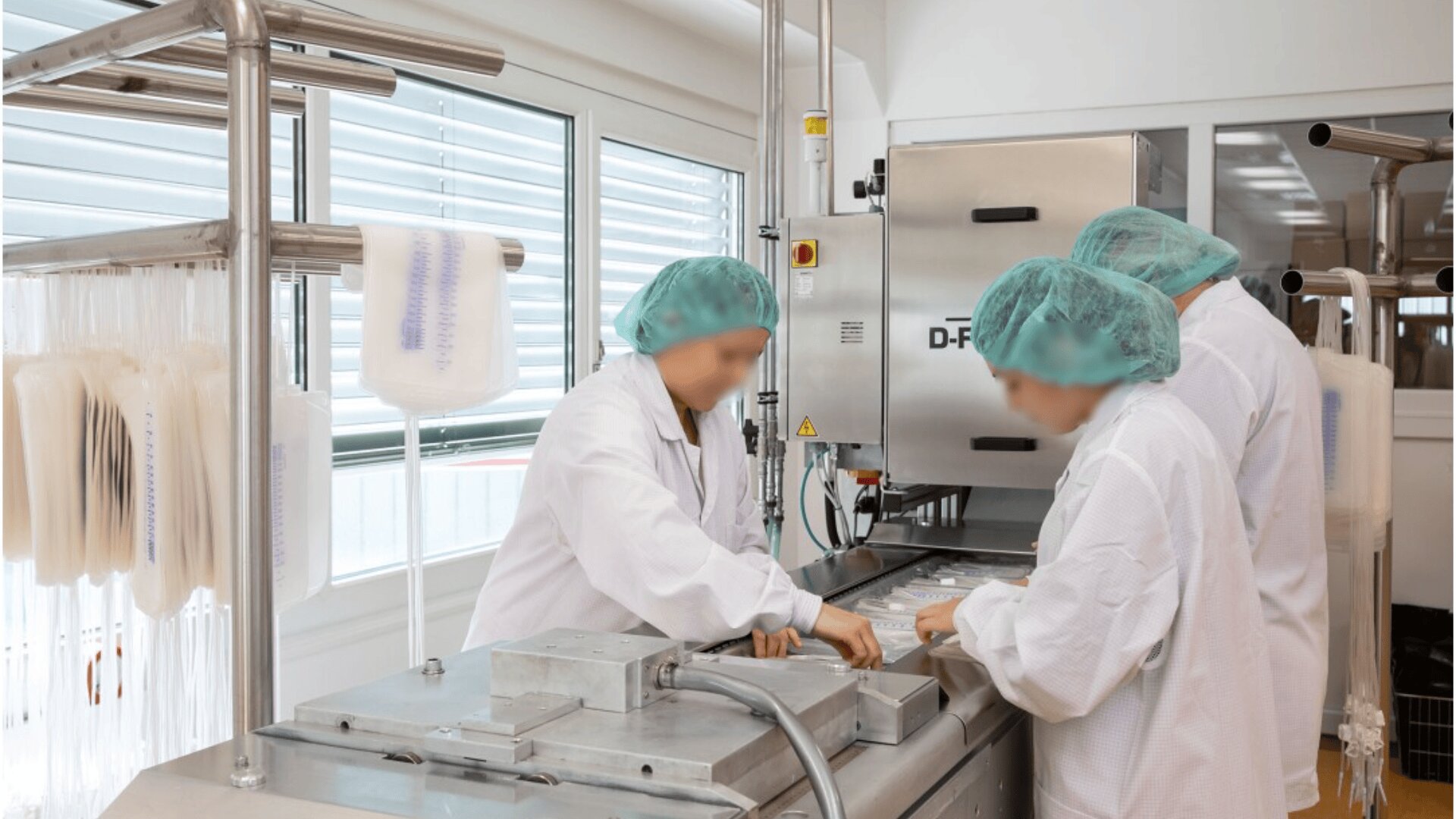 Three people in white lab coats and green hairnets operate packaging equipment in a clean, bright laboratory. Empty plastic bags hang on racks nearby, and machinery is visible in the background.