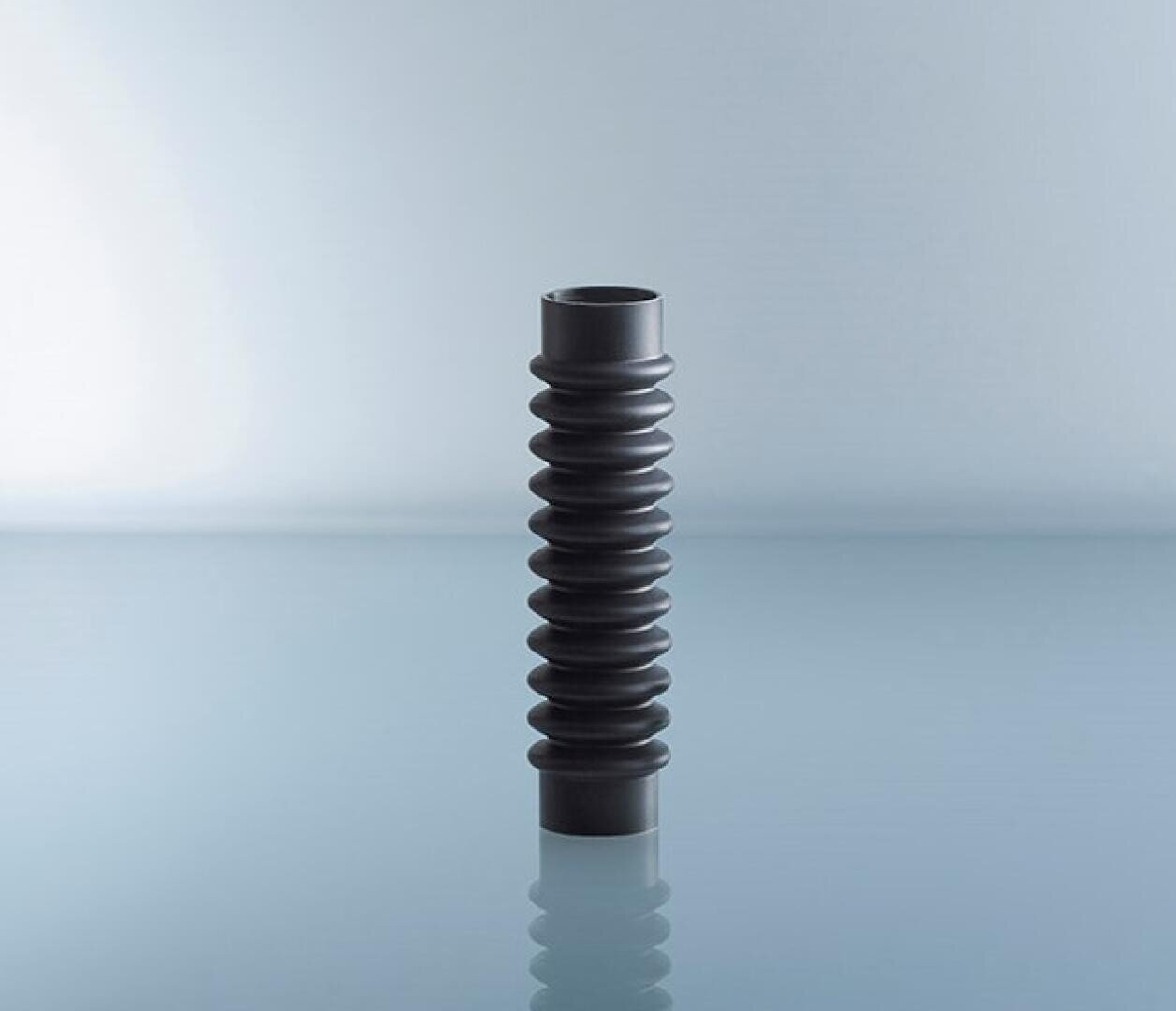 A black, cylindrical, accordion-like rubber bellows stands upright on a shiny, light blue surface with a pale blue color gradient in the background.