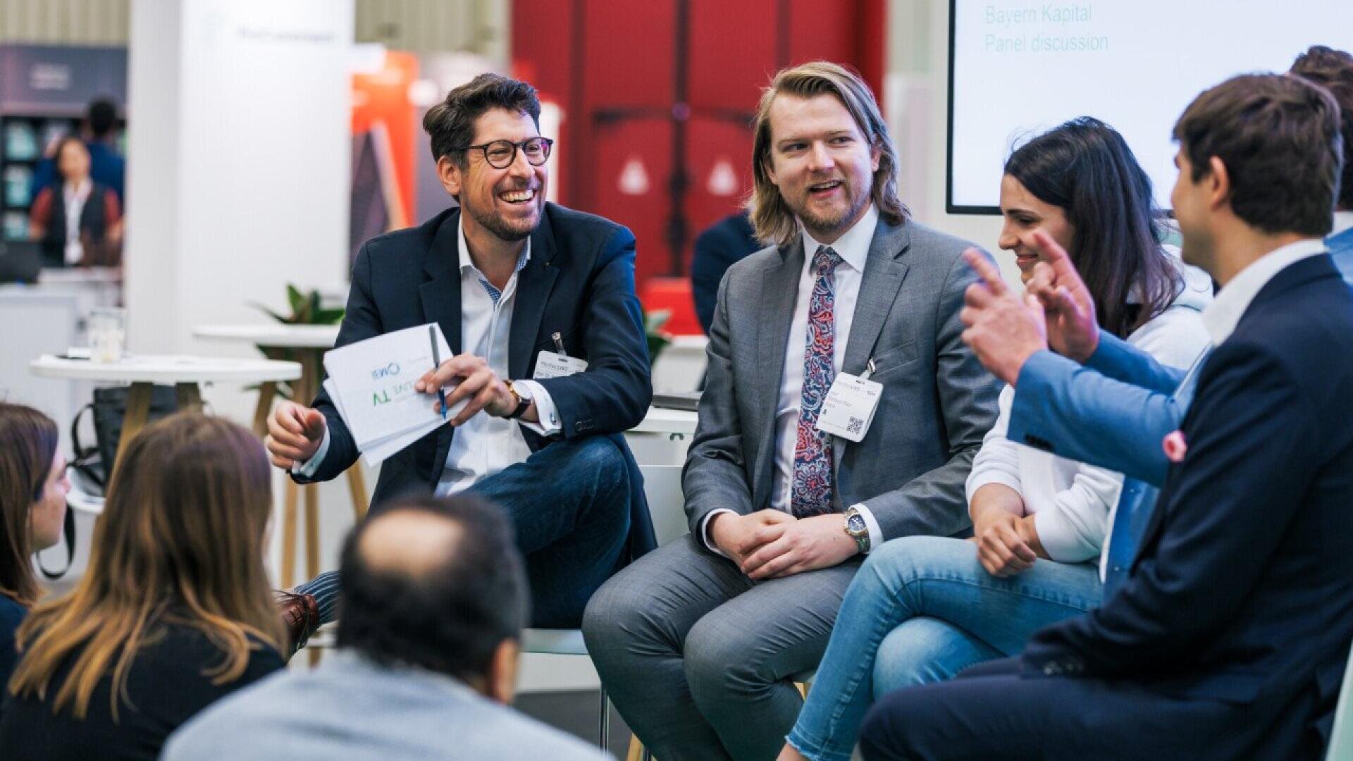 A group of professionally dressed people sit in a semi-circle at an indoor event, smiling and engaging in lively conversation during a panel discussion. One man is holding a notepad and appears to be leading the discussion.