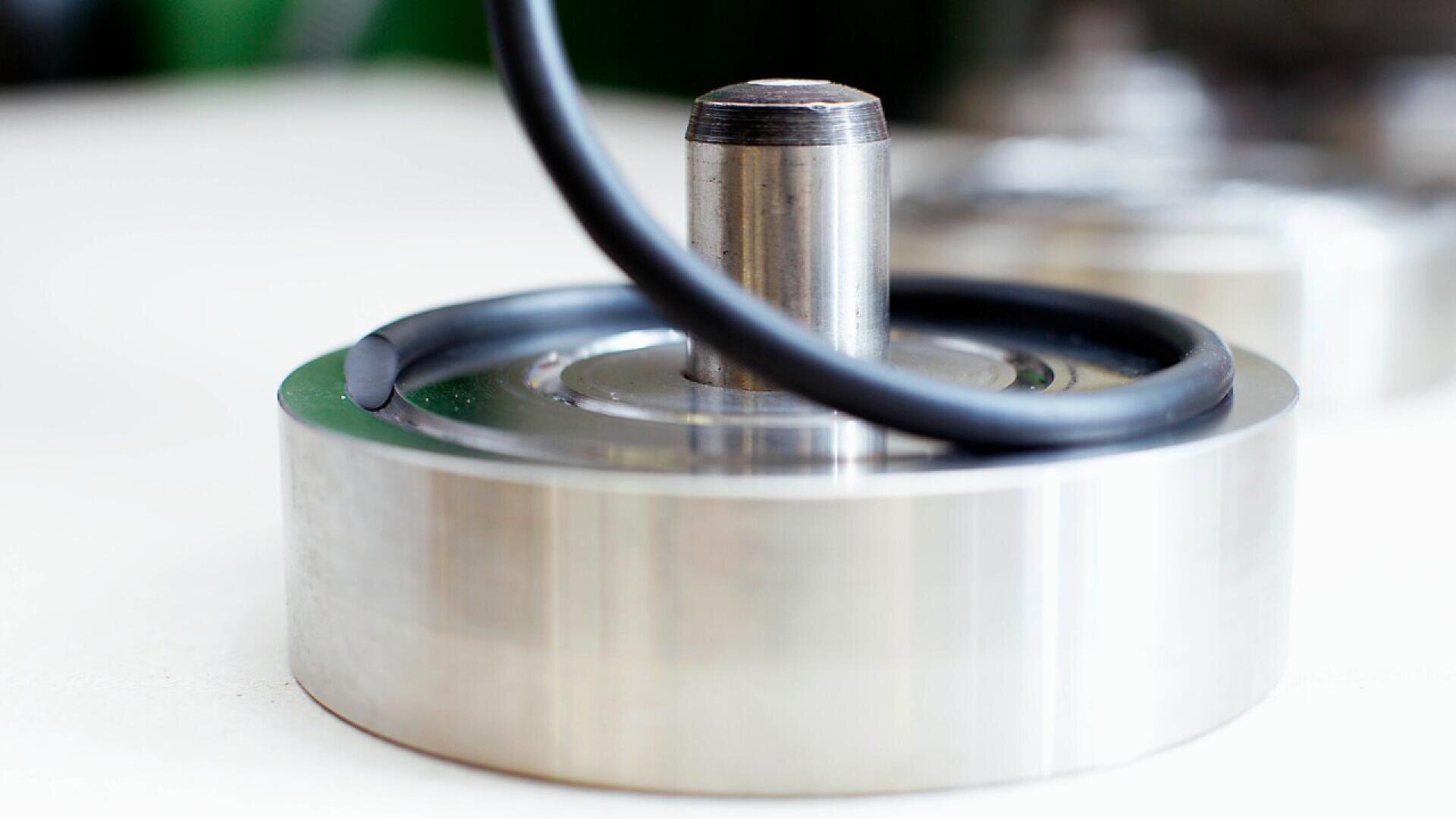 A close-up of a round metallic object with a central cylindrical post. A black cable or hose is looped around the post, resting on the smooth, shiny metal surface. The background is softly blurred.