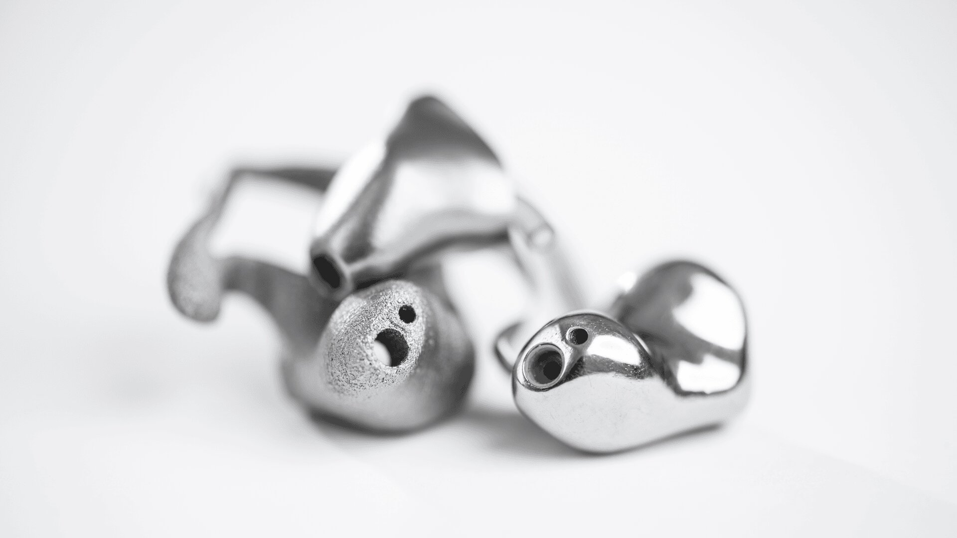 Eine Nahaufnahme von zwei glänzenden, metallischen, individuell geformten Ohrhörern mit kleinen Öffnungen, die auf einer weißen Oberfläche aneinanderliegen.