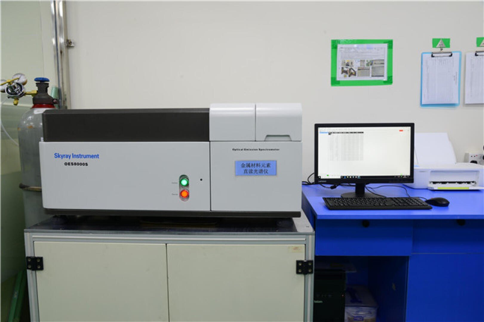 A laboratory setup with a Skyray Instrument OES8000S spectrometer on a cabinet next to a computer on a blue desk, displaying data on the monitor, and laboratory papers on the wall.
