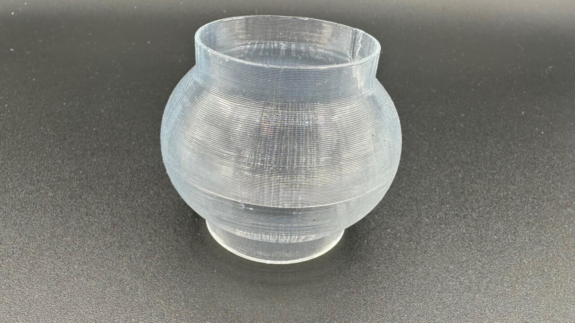A translucent, round, 3D-printed plastic vase with visible horizontal print lines sits on a flat, dark surface.