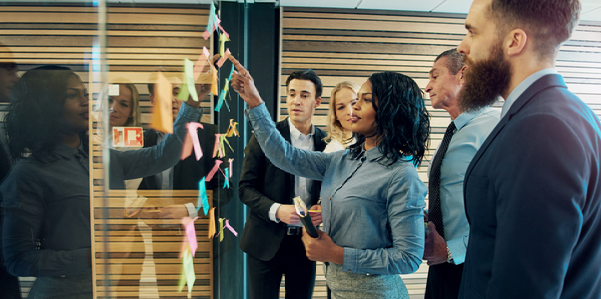 Five business people stand in front of a glass wall covered with colorful sticky notes. A woman points to the notes while the others watch attentively, suggesting a collaborative planning or brainstorming session.