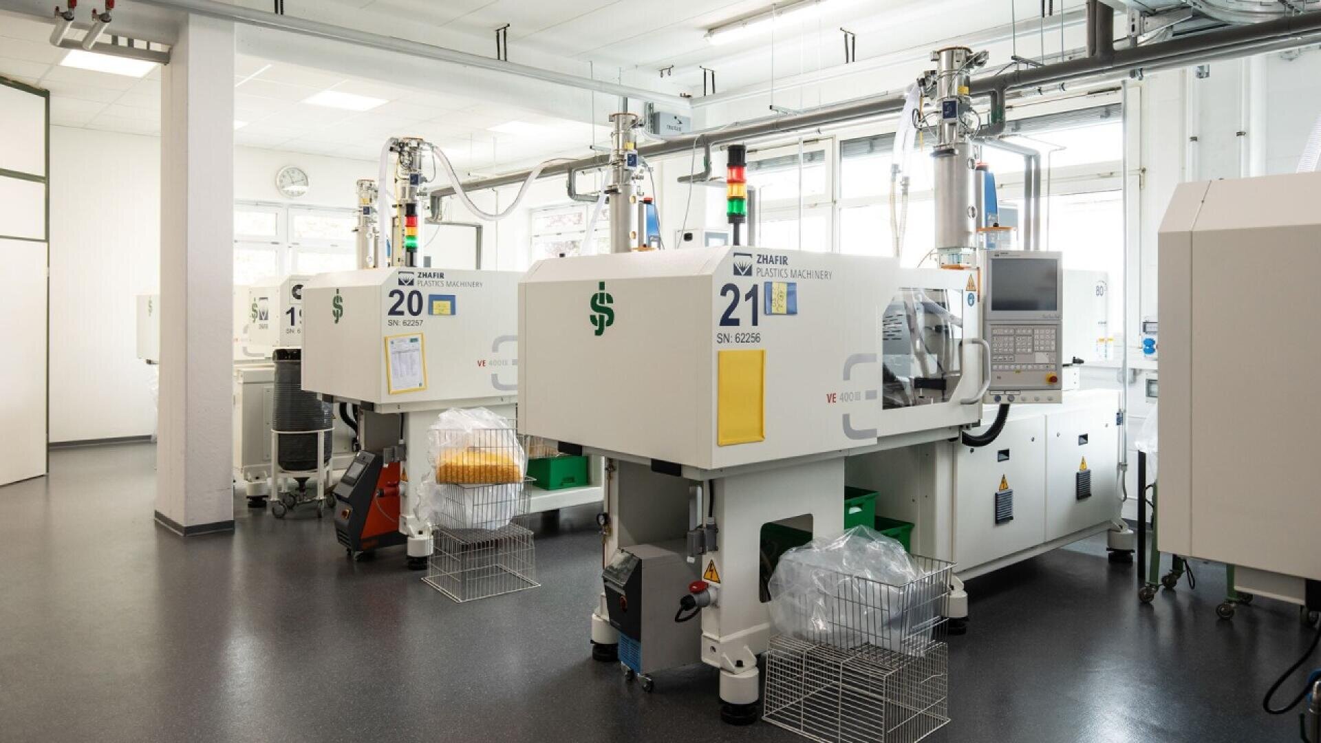 Two modern injection molding machines with control panels and numbered labels are positioned in a bright, clean factory setting. Wire baskets and storage bins are placed underneath the machines.