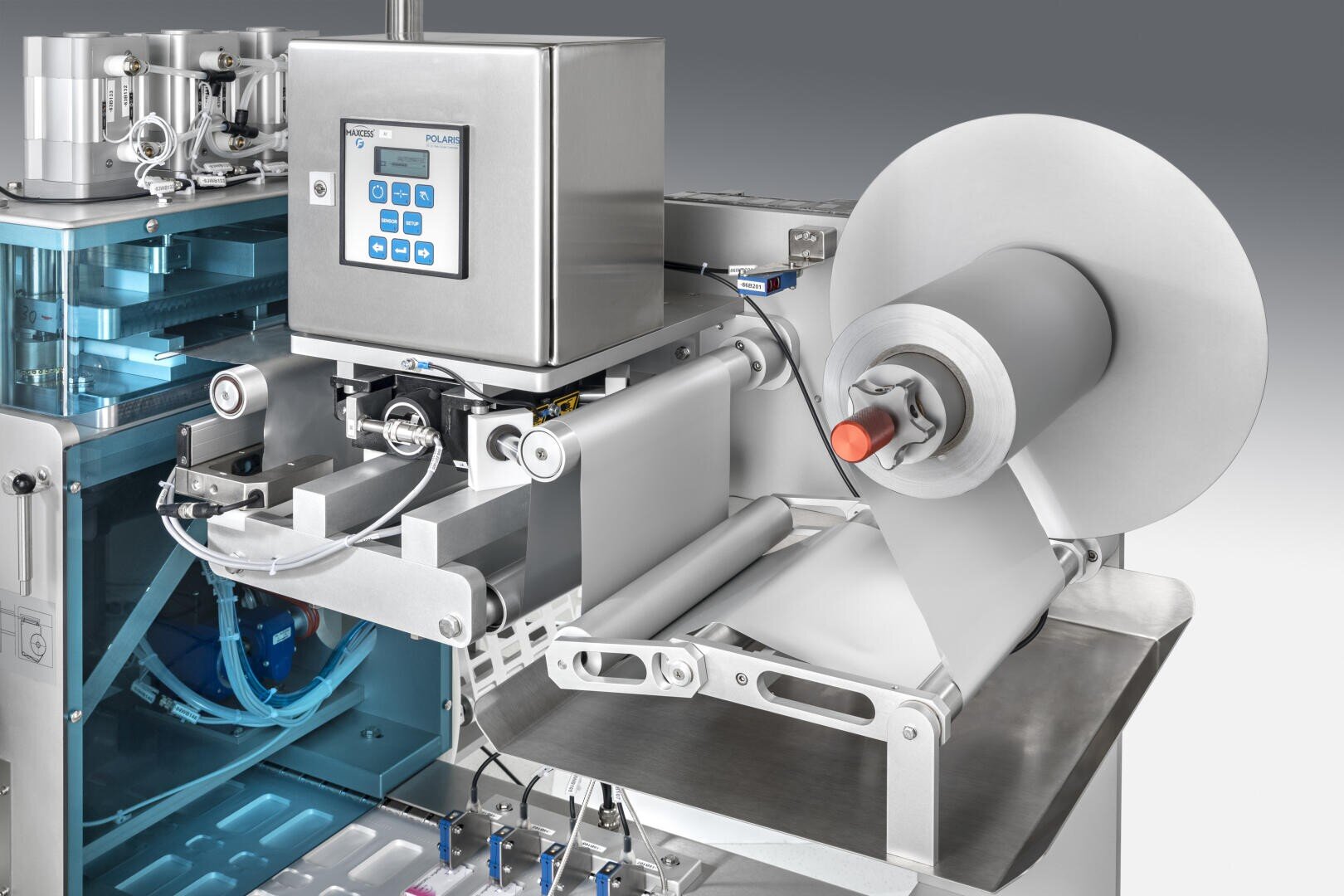 Nahaufnahme einer automatisierten Verpackungsmaschine mit einer Rolle Folienmaterial, einem Bedienfeld mit Knöpfen und verschiedenen metallischen Komponenten zum Versiegeln und Verarbeiten von Produkten.