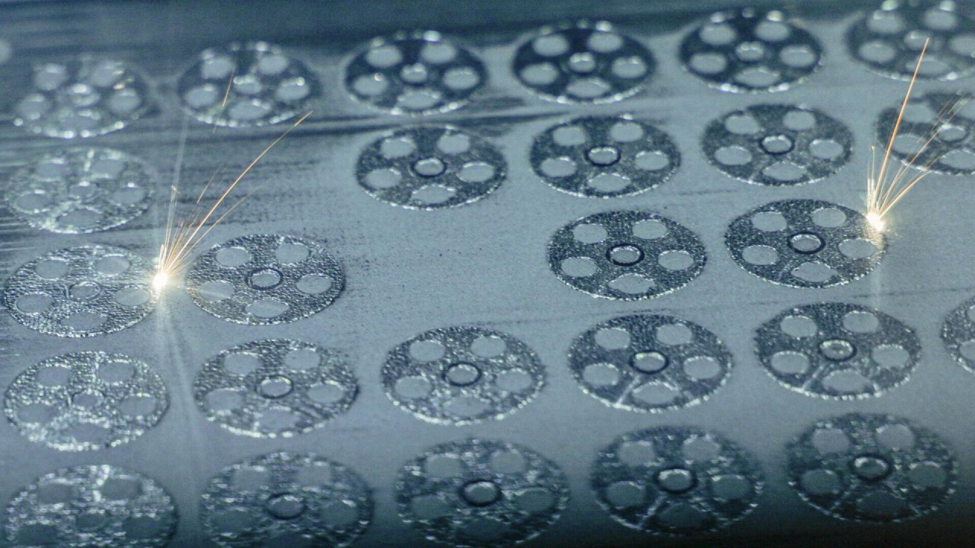 Close-up of a metal 3D printing process showing a grid of circular patterns with two laser beams creating bright sparks on the metallic surface.