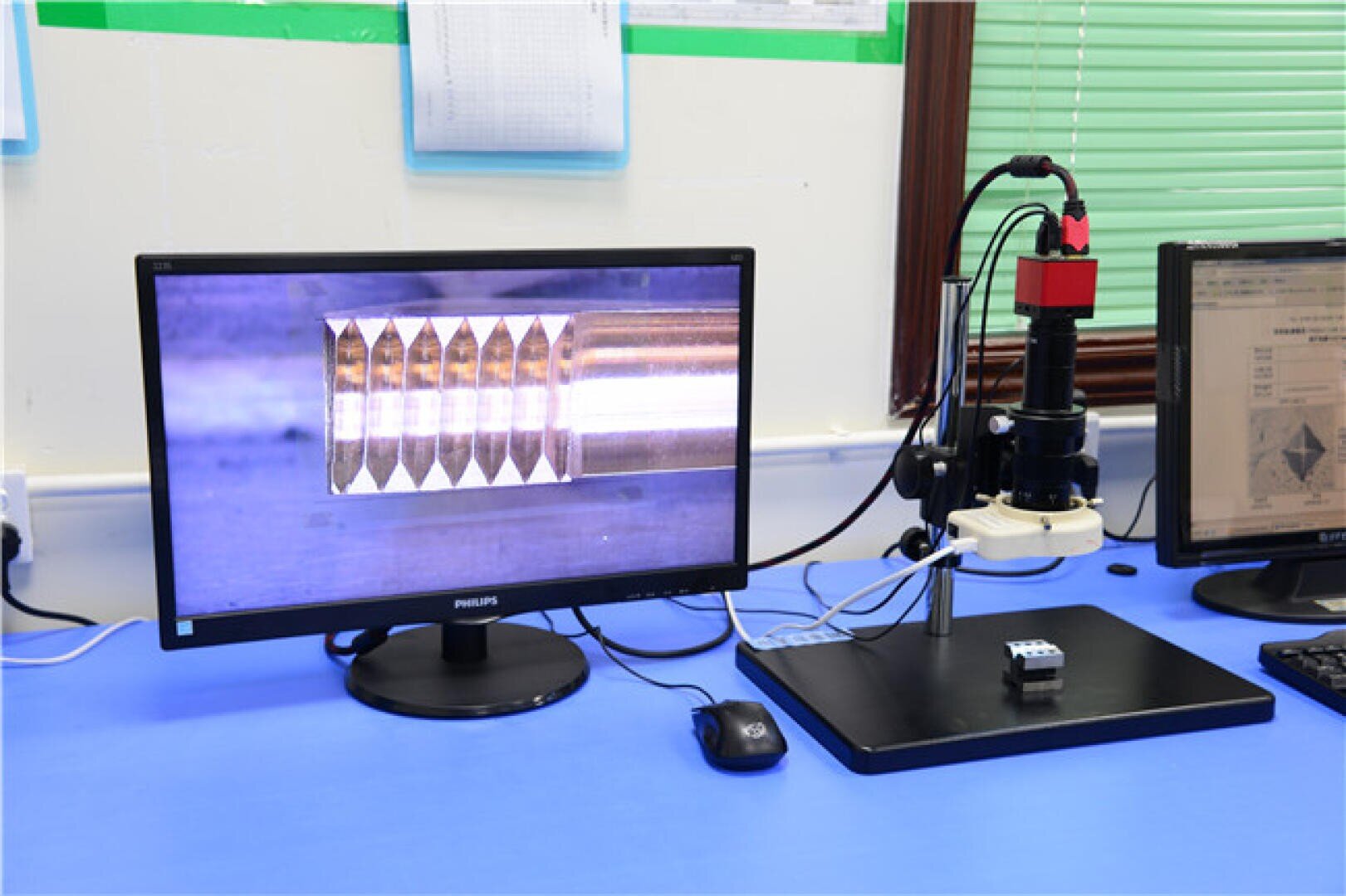 A computer monitor displays a magnified view of a metallic object with grooves. Next to it, a digital microscope is positioned over a small metal sample on a blue desk in a laboratory setting.