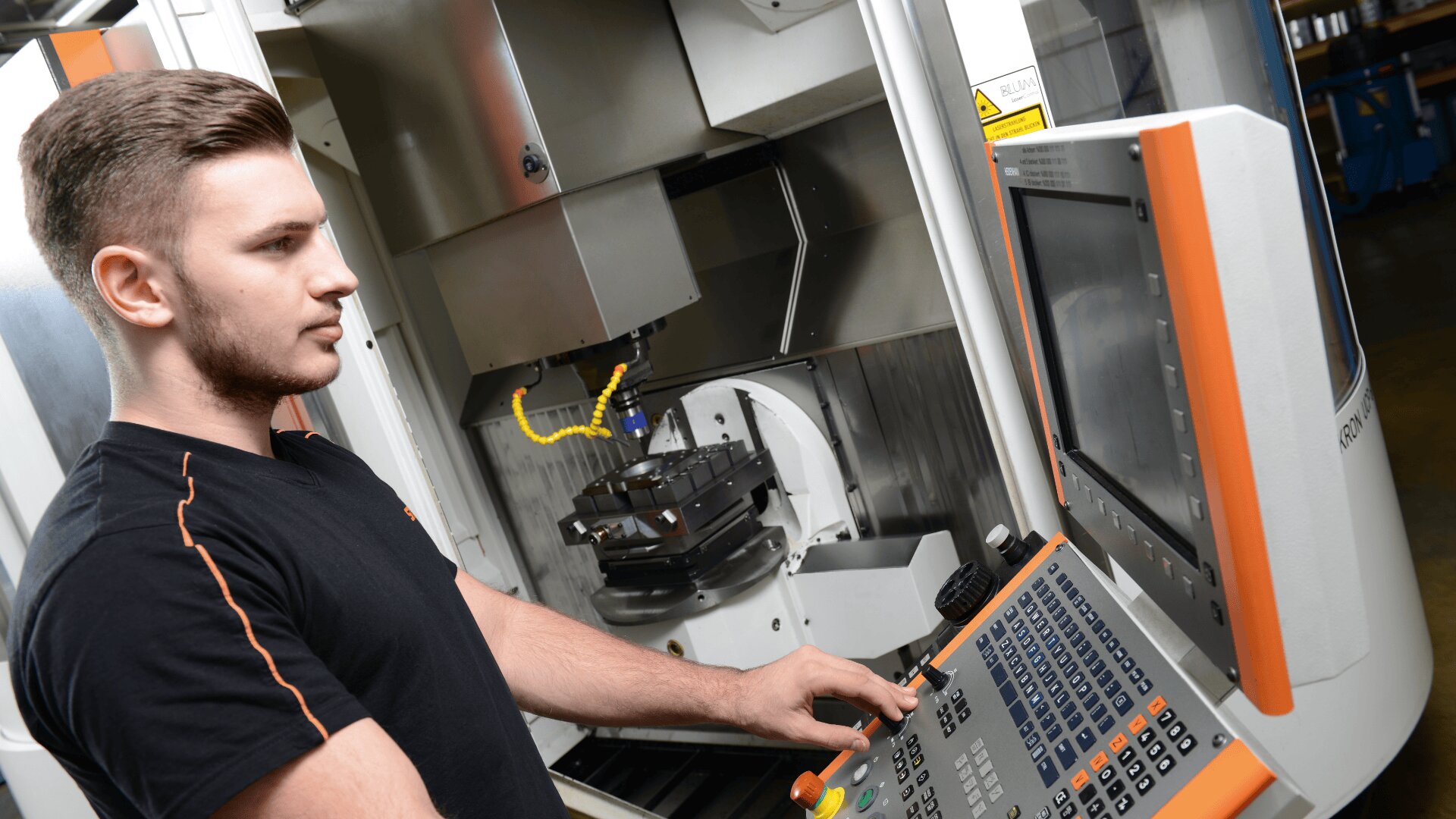 Ein Mann bedient eine CNC-Maschine mit einem Bedienfeld mit Tasten und einem Bildschirm in einer Industrieumgebung. In der Maschine befindet sich ein Metallwerkstück, und es sind gelbe Kabel zu sehen.