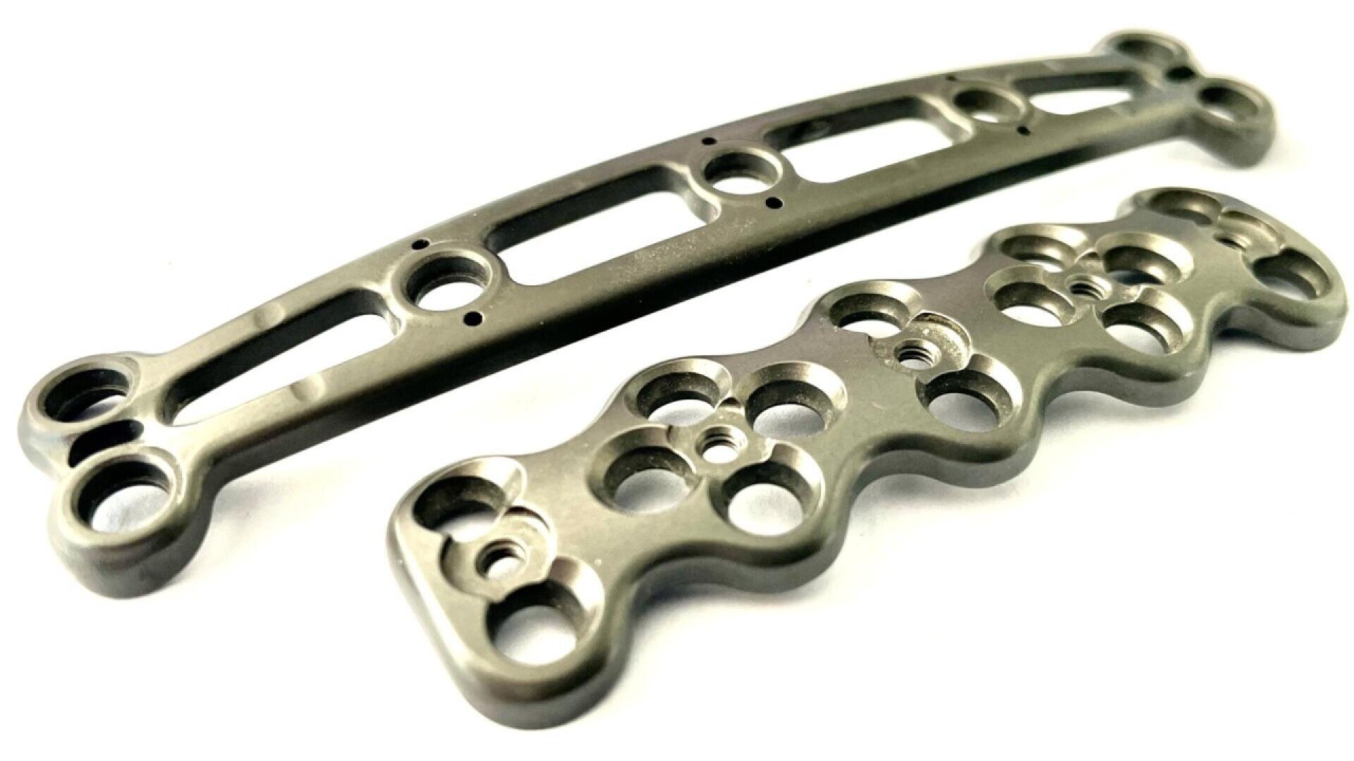 Two metallic orthopedic bone plates with multiple holes and curved edges, designed for surgical bone fixation, are placed on a white background.