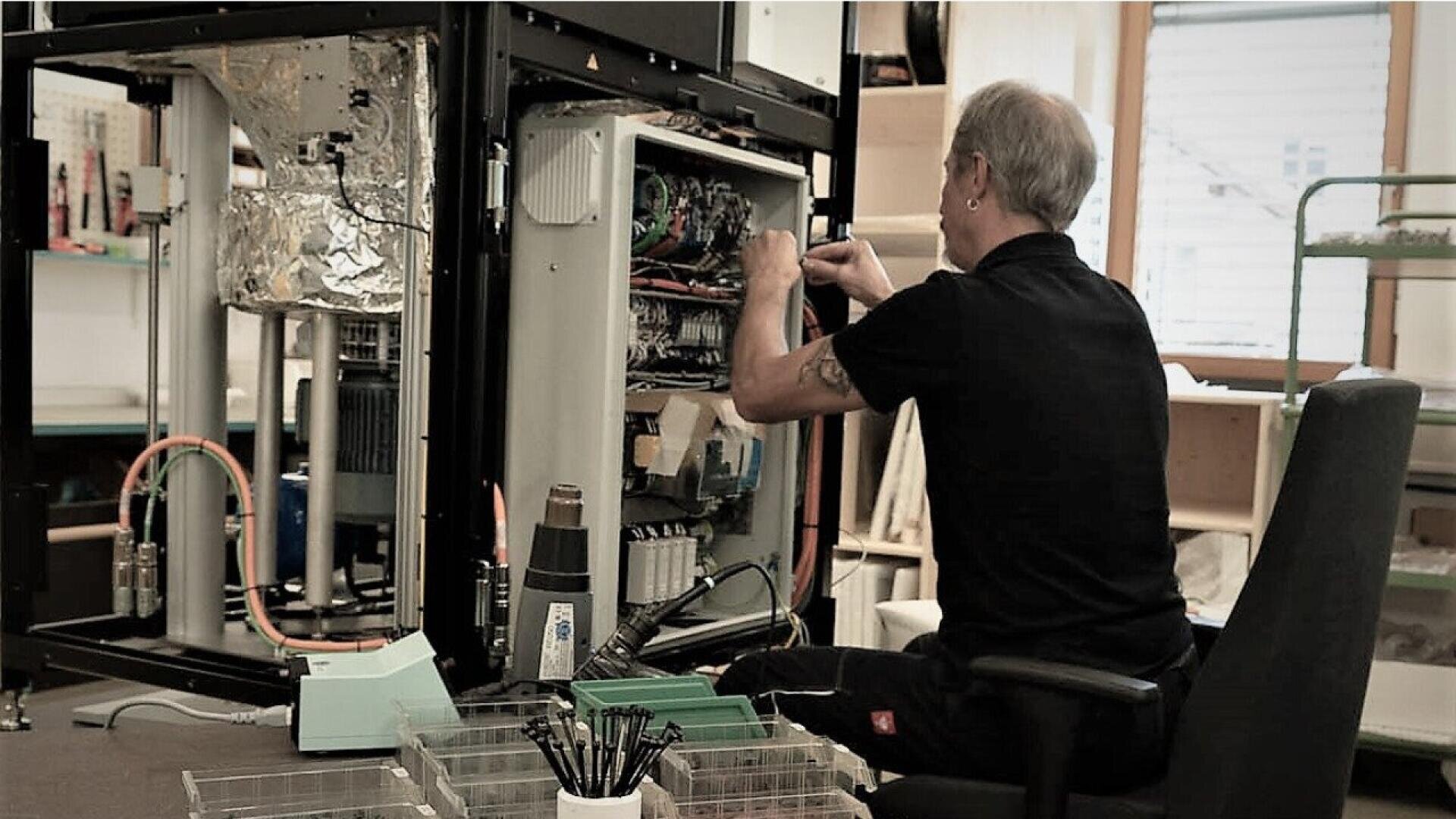 A man sits in a chair repairing or assembling electronic equipment inside a large open cabinet, with tools and parts organized in containers nearby.