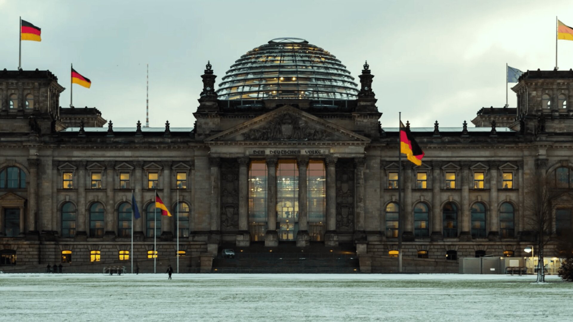Das Reichstagsgebäude in Berlin, Deutschland, mit seiner Glaskuppel und den wehenden deutschen Fahnen, ist an einem bewölkten Tag zu sehen, vor dem etwas Schnee auf dem Boden liegt.