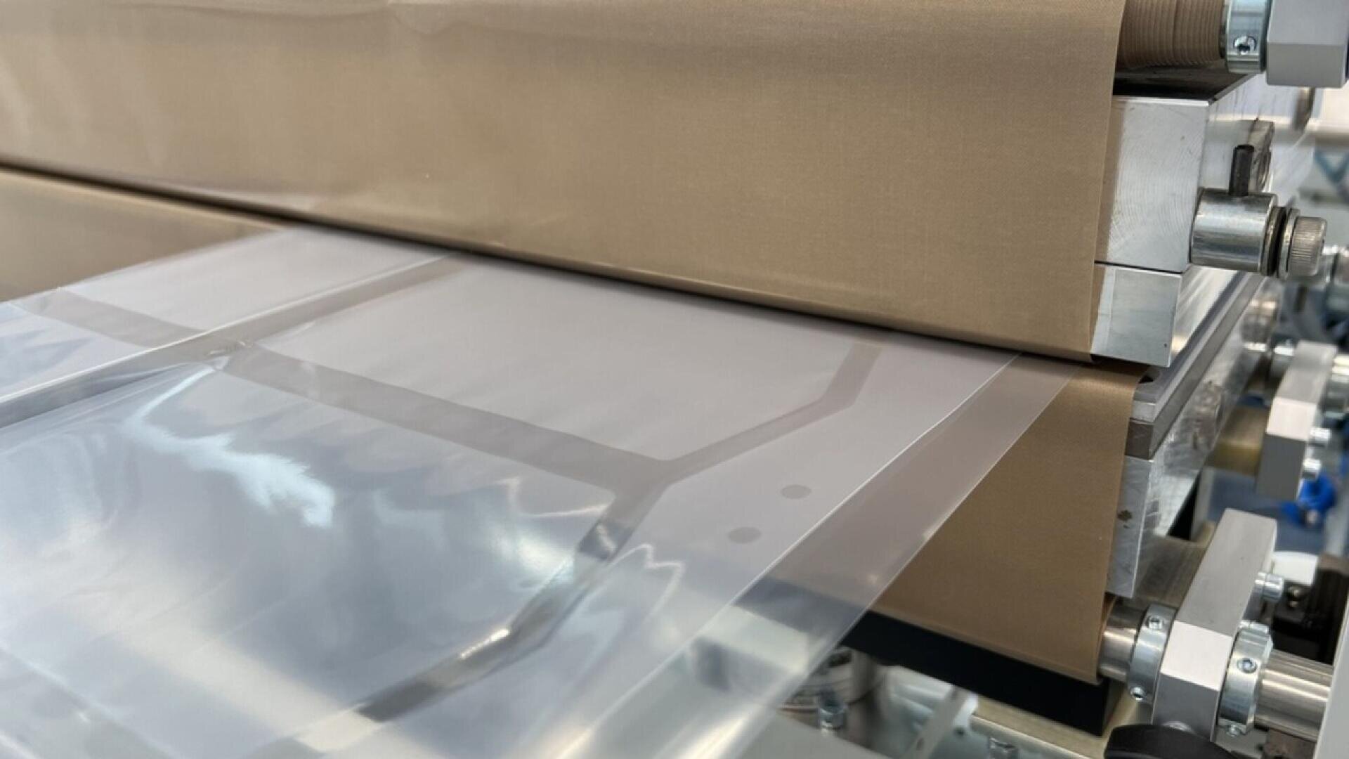 A close-up of a machine processing and pressing transparent plastic film between two rollers in a manufacturing setting.