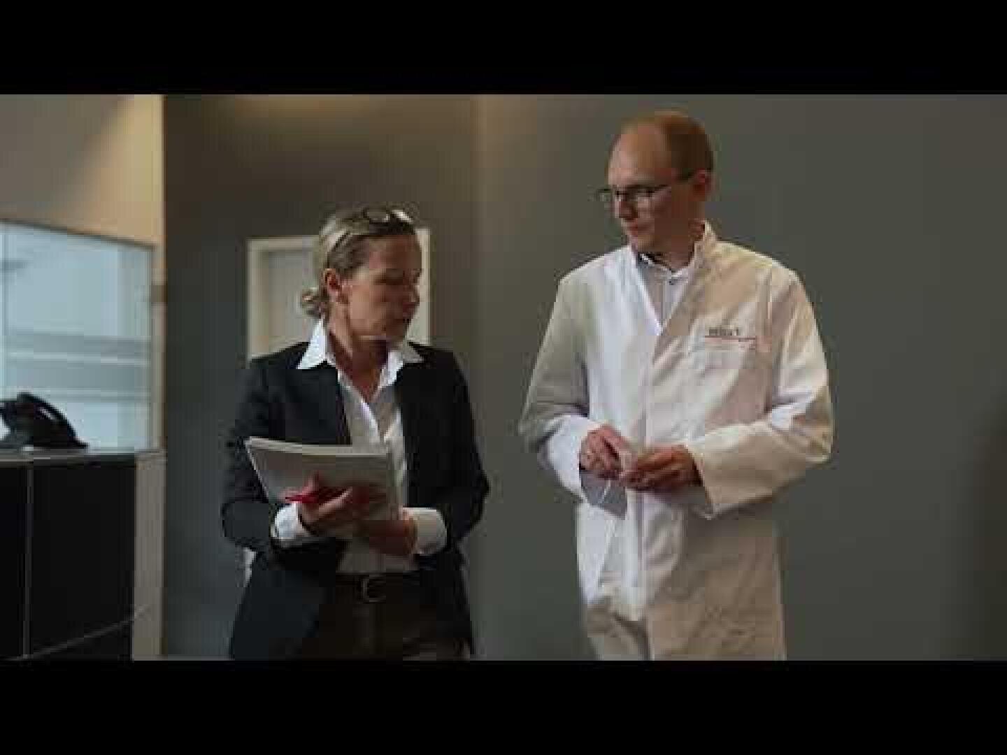 Eine Frau in Geschäftskleidung, die Papiere in der Hand hält, und ein Mann in einem weißen Laborkittel unterhalten sich beim Gang durch ein Büro oder eine medizinische Einrichtung.